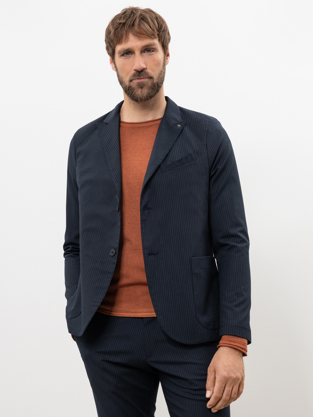 Ein Mann mit kurzen braunen Haaren und einem Bart trägt einen marineblauen Nadelstreifenanzug über einem rostfarbenen Pullover. Er steht vor einem schlichten weißen Hintergrund, hat die Hände in den Taschen und schaut in die Kamera.