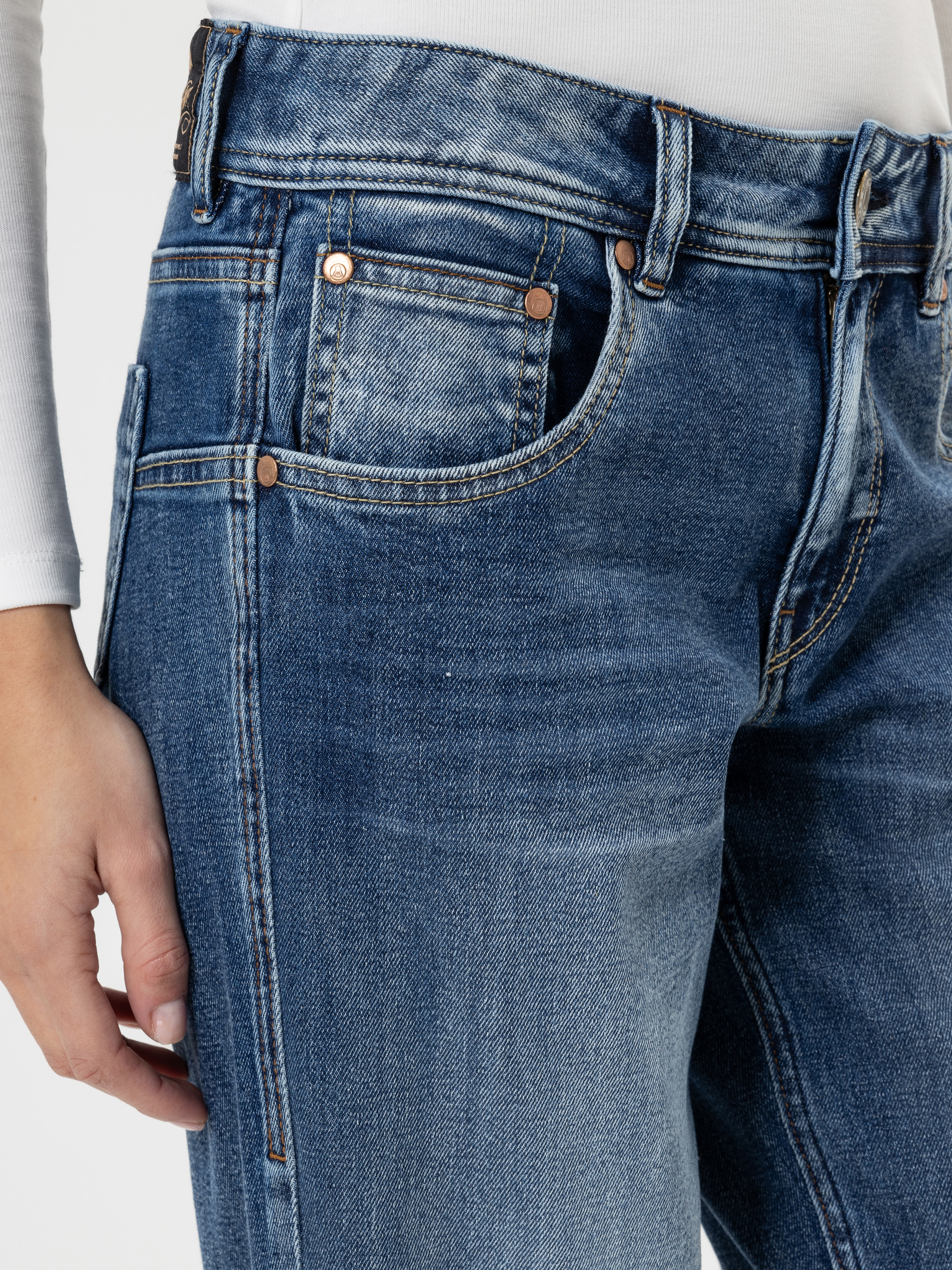 Eine Nahaufnahme einer Person, die eine blaue Jeans und ein weißes langärmeliges Oberteil trägt, wobei die linke Hand entspannt an der Seite liegt. Der Fokus liegt auf der Jeanstasche und den Nähten.