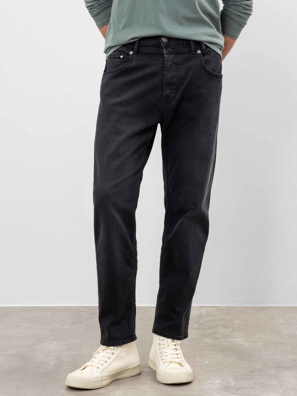 Eine Person in schwarzen Jeans, einem hellgrünen langärmeligen Hemd mit hochgekrempelten Ärmeln und cremefarbenen Turnschuhen mit hohen Absätzen steht auf einem Betonboden vor einer schlichten hellgrauen Wand.