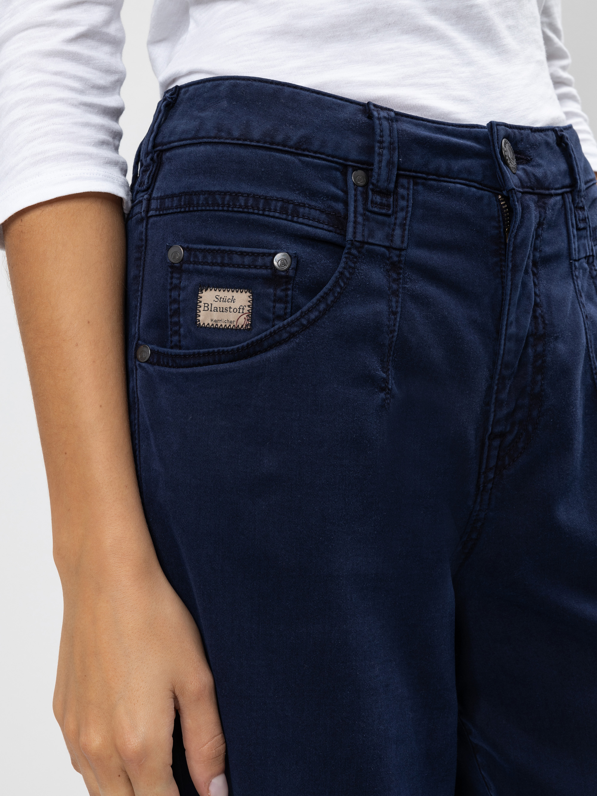 Eine Person trägt eine dunkelblaue Jeans und ein weißes langärmeliges Hemd. Das Foto konzentriert sich auf den oberen Teil der Jeans und zeigt ein kleines Etikett auf der Münztasche.