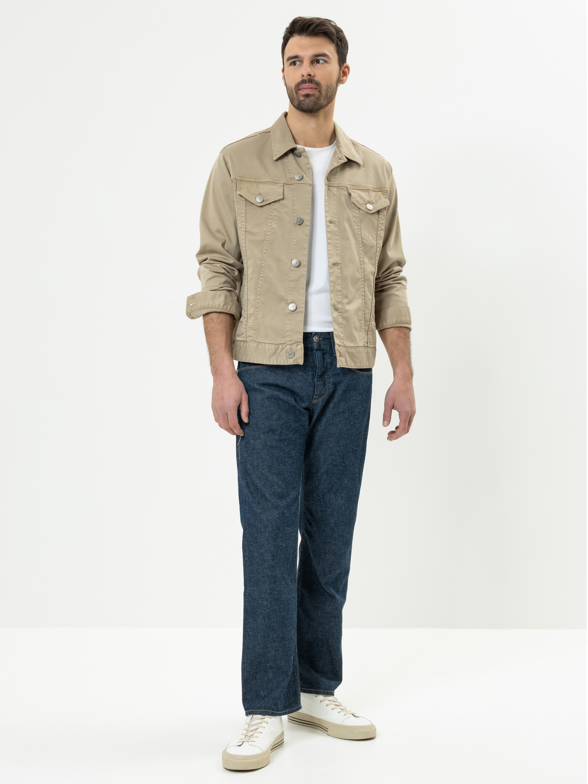 Ein Mann, der eine beige Jeansjacke über einem weißen T-Shirt, blaue Jeans und weiße Turnschuhe trägt, steht vor einem schlichten weißen Hintergrund.