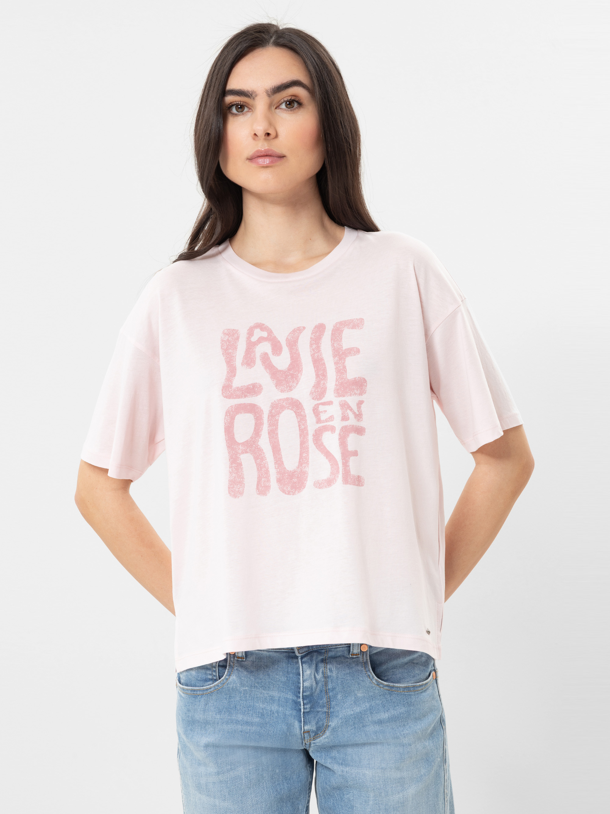 Eine Frau mit langen dunklen Haaren trägt ein hellrosa T-Shirt mit der Aufschrift La Vie en Rose in verblassten rosafarbenen Buchstaben und hellblaue Jeans vor einem schlichten weißen Hintergrund.