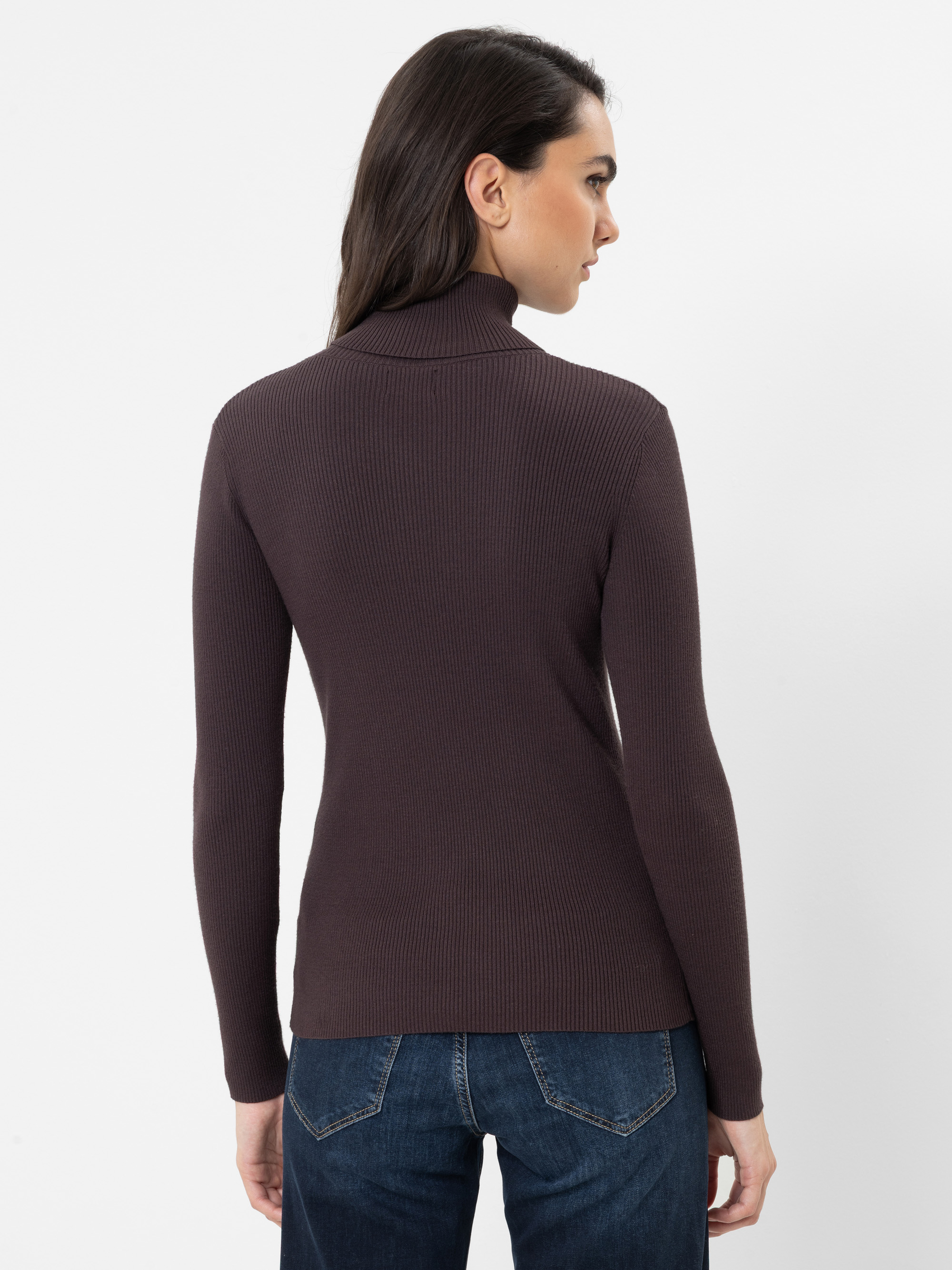 Eine Frau mit langen braunen Haaren, die einen dunkelbraunen, gerippten Rollkragenpullover und blaue Jeans trägt und mit dem Rücken zur Kamera vor einem schlichten weißen Hintergrund steht.