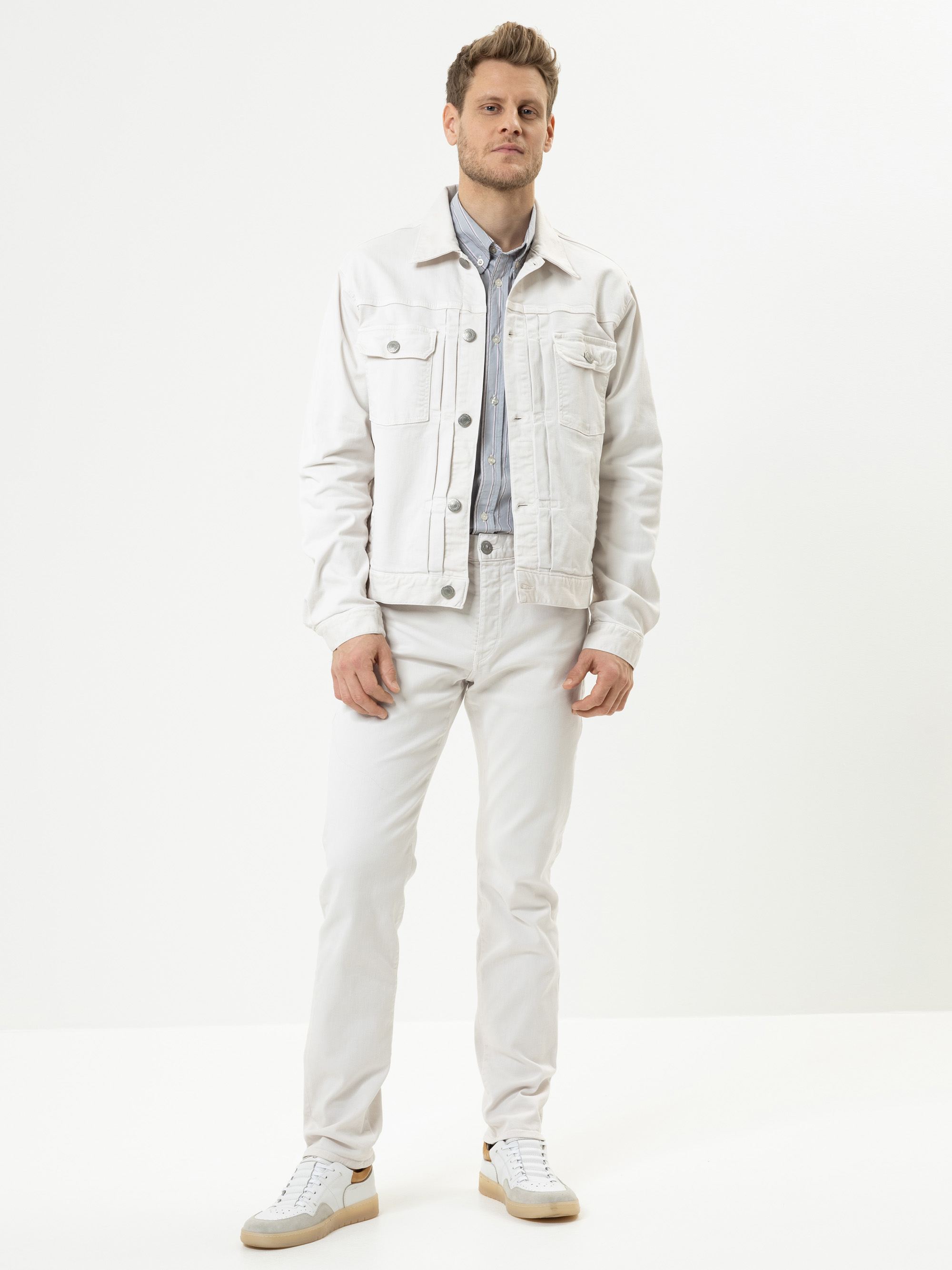 Ein Mann steht vor einem schlichten weißen Hintergrund und trägt eine helle Jeansjacke, ein hellblaues Button-up-Hemd, eine weiße Hose und weiße Turnschuhe. Er hat kurze Haare und einen neutralen Gesichtsausdruck.