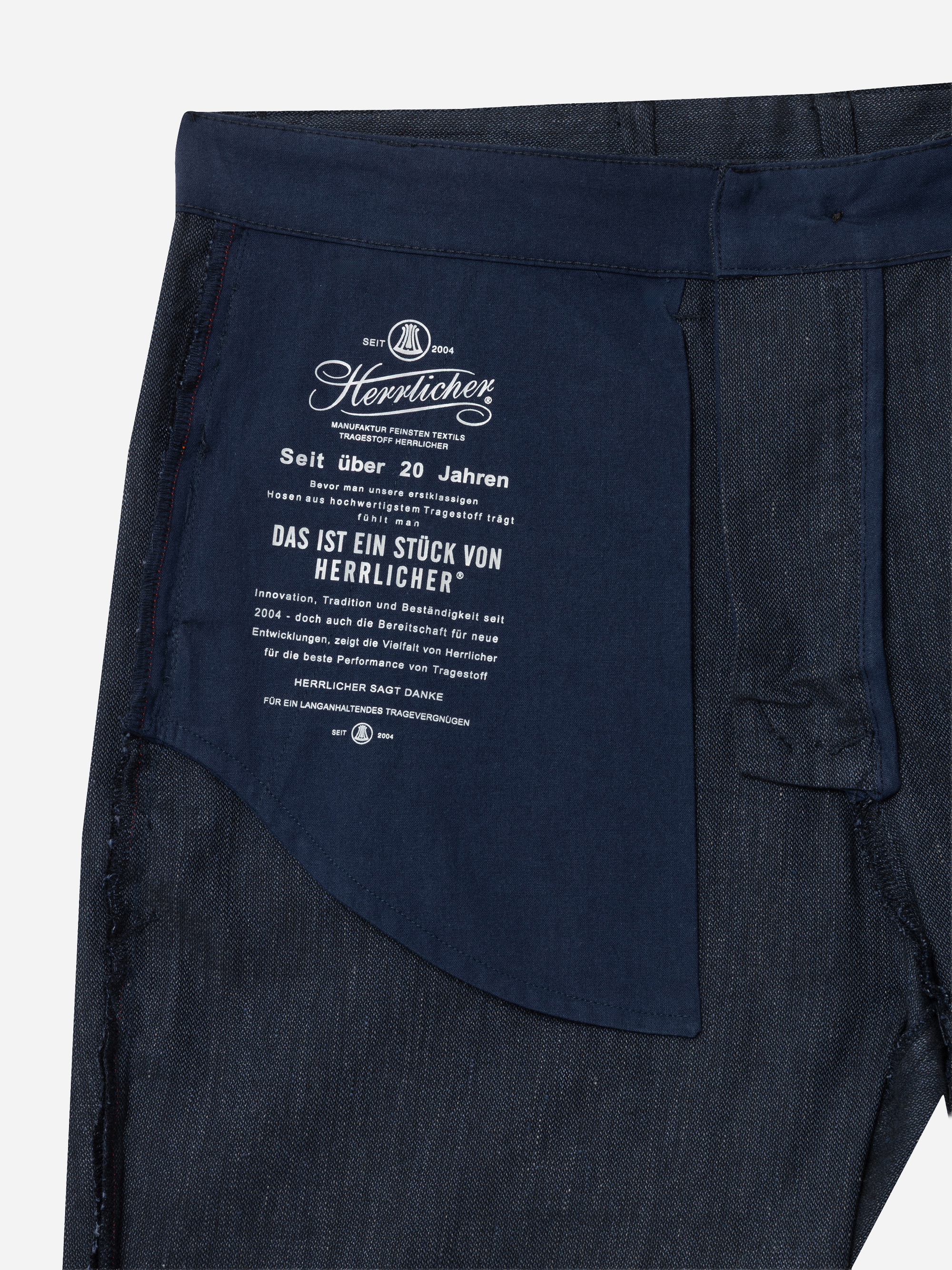 Nahaufnahme der vorderen Innentasche einer dunklen Denim-Jeans mit weiß gedrucktem Branding und Text von Herrlicher in deutscher Sprache, der die Qualität und das Erbe der Marke hervorhebt.