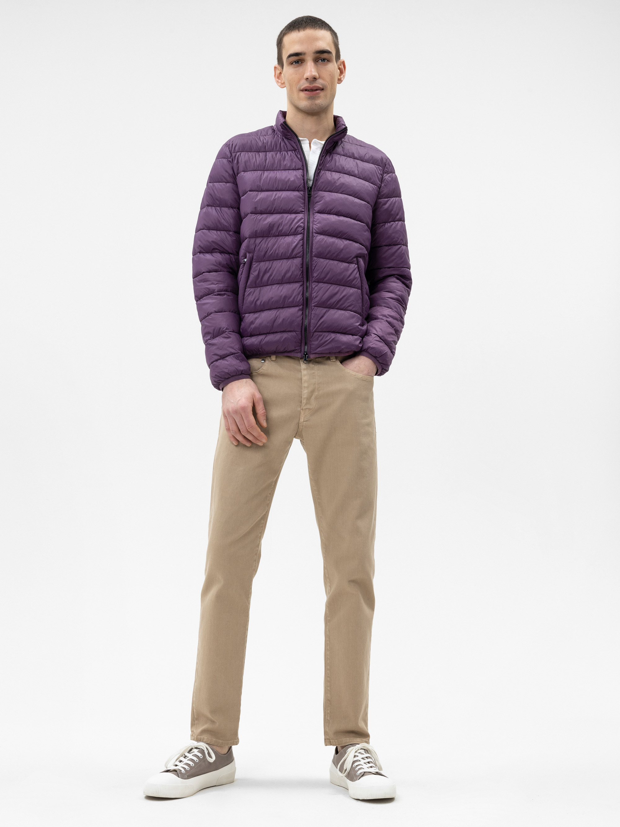 Ein Mann steht mit dem Gesicht nach vorne und trägt eine lila Pufferjacke, eine beige Hose und weiße Turnschuhe. Er hat kurzes Haar und seine Hände stecken in den Jackentaschen, vor einem schlichten weißen Hintergrund.