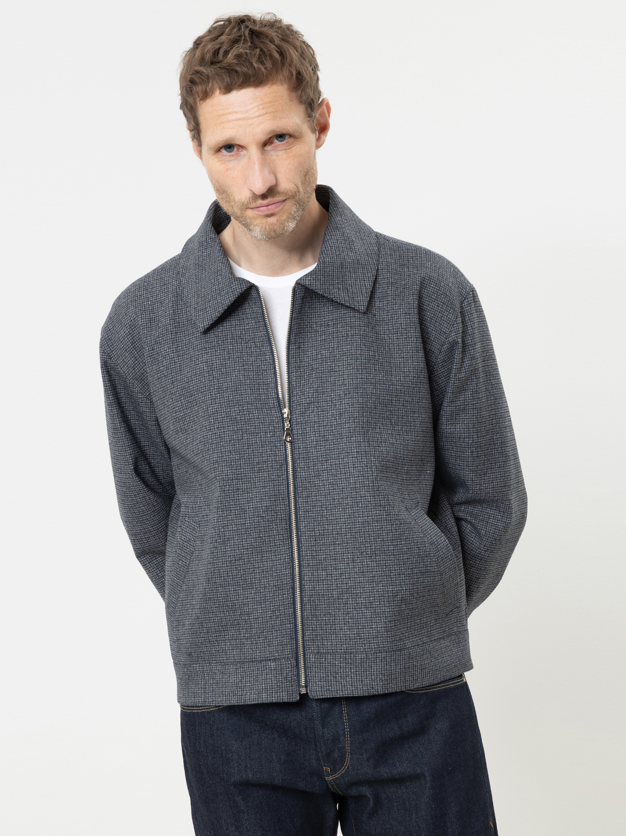 Ein Mann mit kurzen braunen Haaren, der eine graue Jacke mit Reißverschluss über einem weißen Hemd und einer dunklen Jeans trägt, steht vor einem schlichten weißen Hintergrund, den Blick leicht nach unten gerichtet und die Hände hinter dem Rücken v