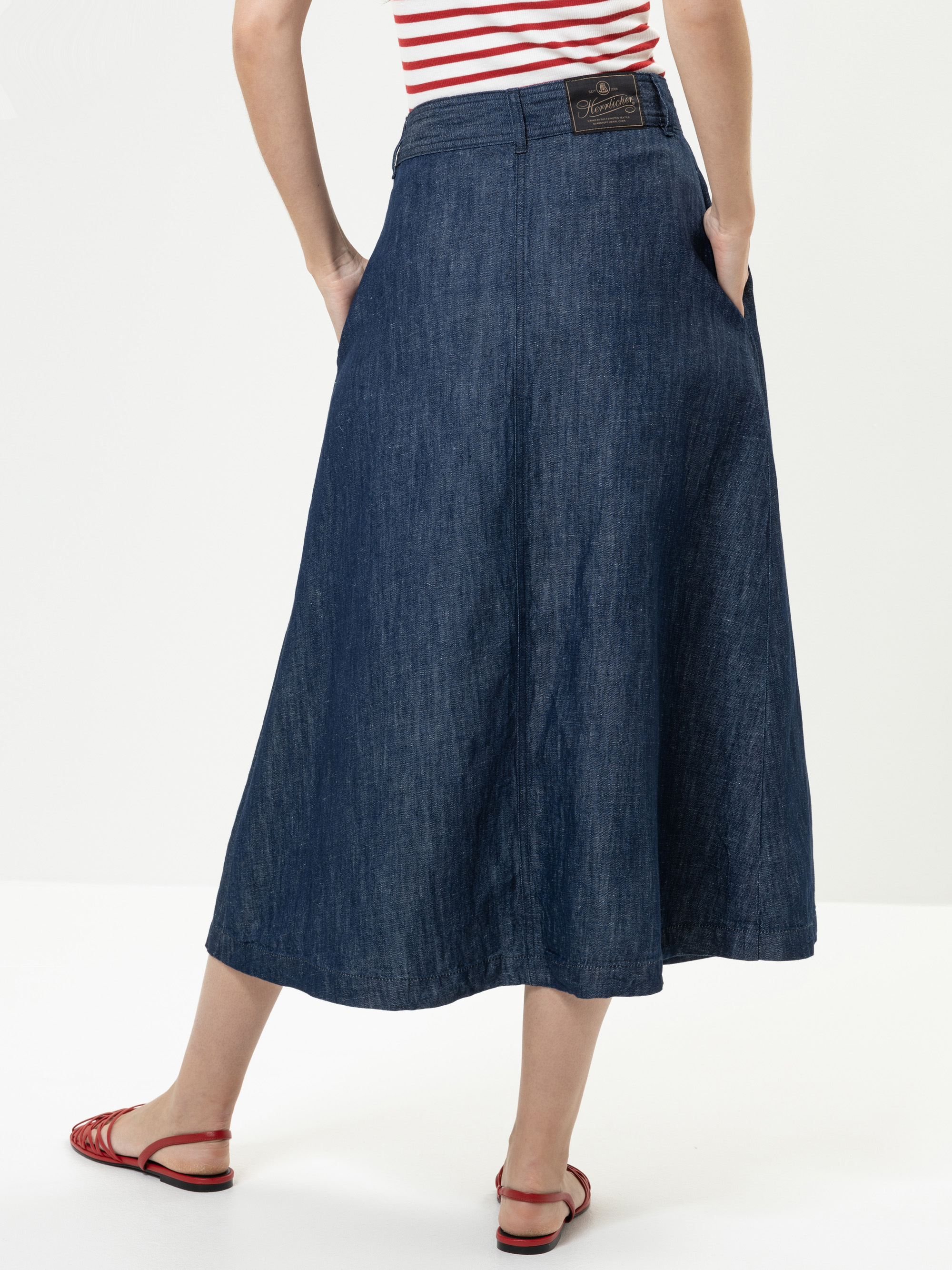 Eine Person, die einen langen, dunklen Jeansrock, rote Sandalen und ein rot-weiß gestreiftes Oberteil trägt, wird von hinten mit den Händen in den Taschen gezeigt. Der Hintergrund ist einfarbig und hell.