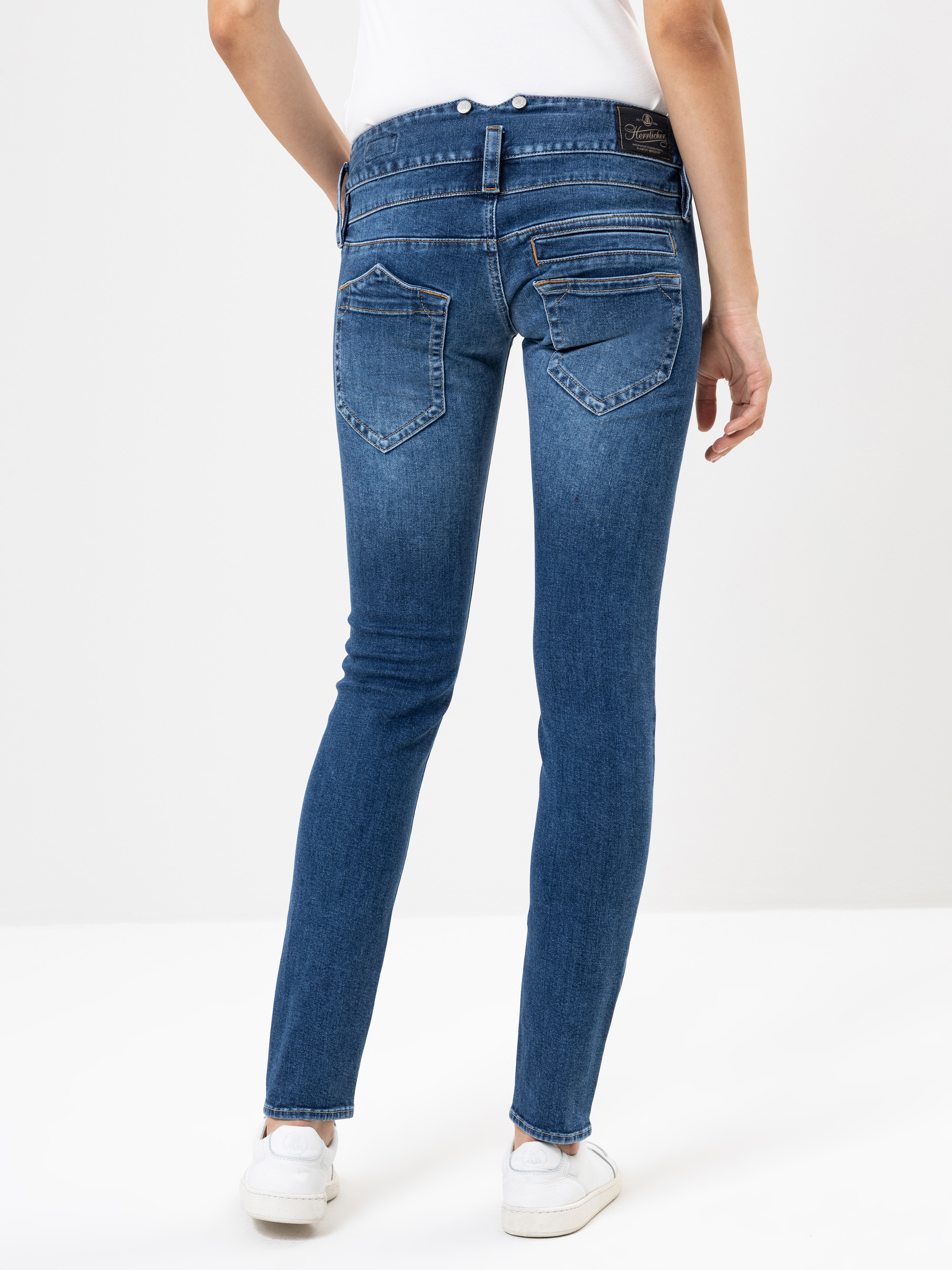 Eine Person, die ein weißes Hemd, blaue Röhrenjeans und weiße Turnschuhe trägt, steht mit dem Rücken zur Kamera vor einem schlichten weißen Hintergrund.