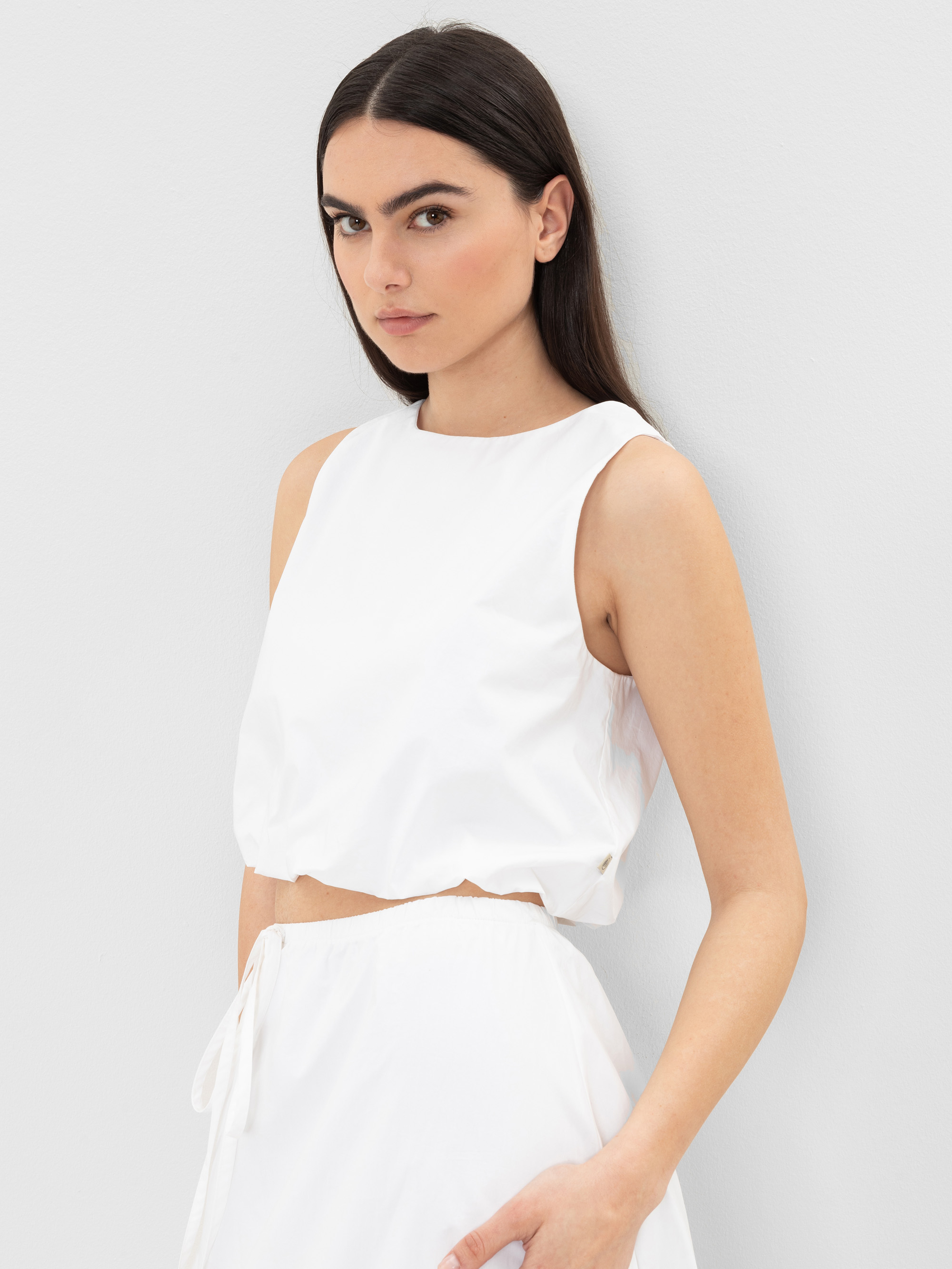 Eine Frau mit langen dunklen Haaren trägt ein ärmelloses weißes Crop-Top und einen dazu passenden weißen Rock. Sie steht vor einer hellgrauen Wand und schaut mit neutralem Blick leicht zur Seite.