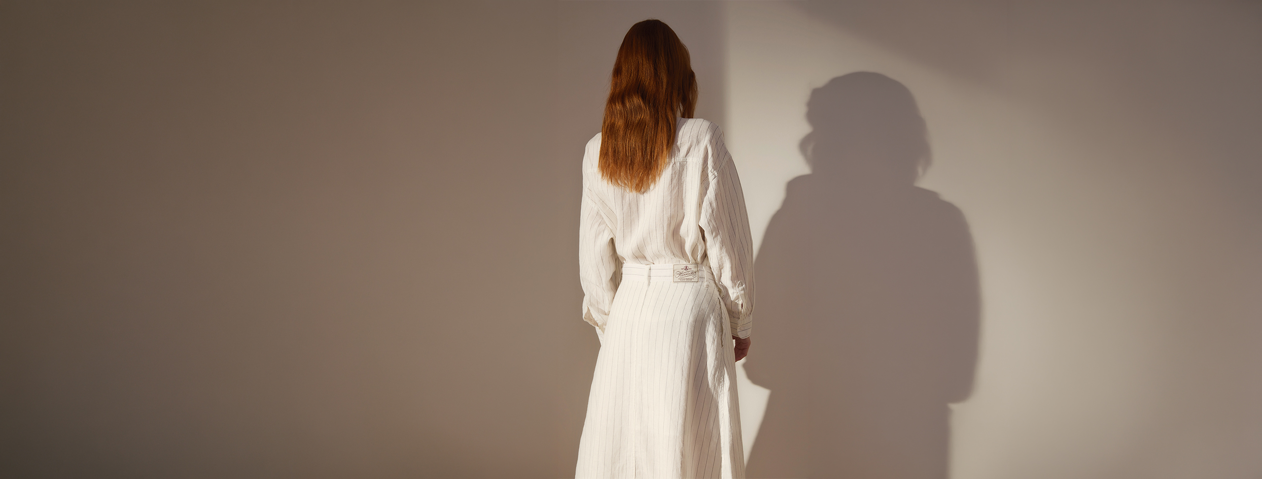 Eine Person mit langen braunen Haaren, die ein langes weißes Kleid trägt, steht mit dem Rücken zur Kamera vor einer beigen Wand. Das Licht wirft einen sanften Schatten der Person auf die Wand.