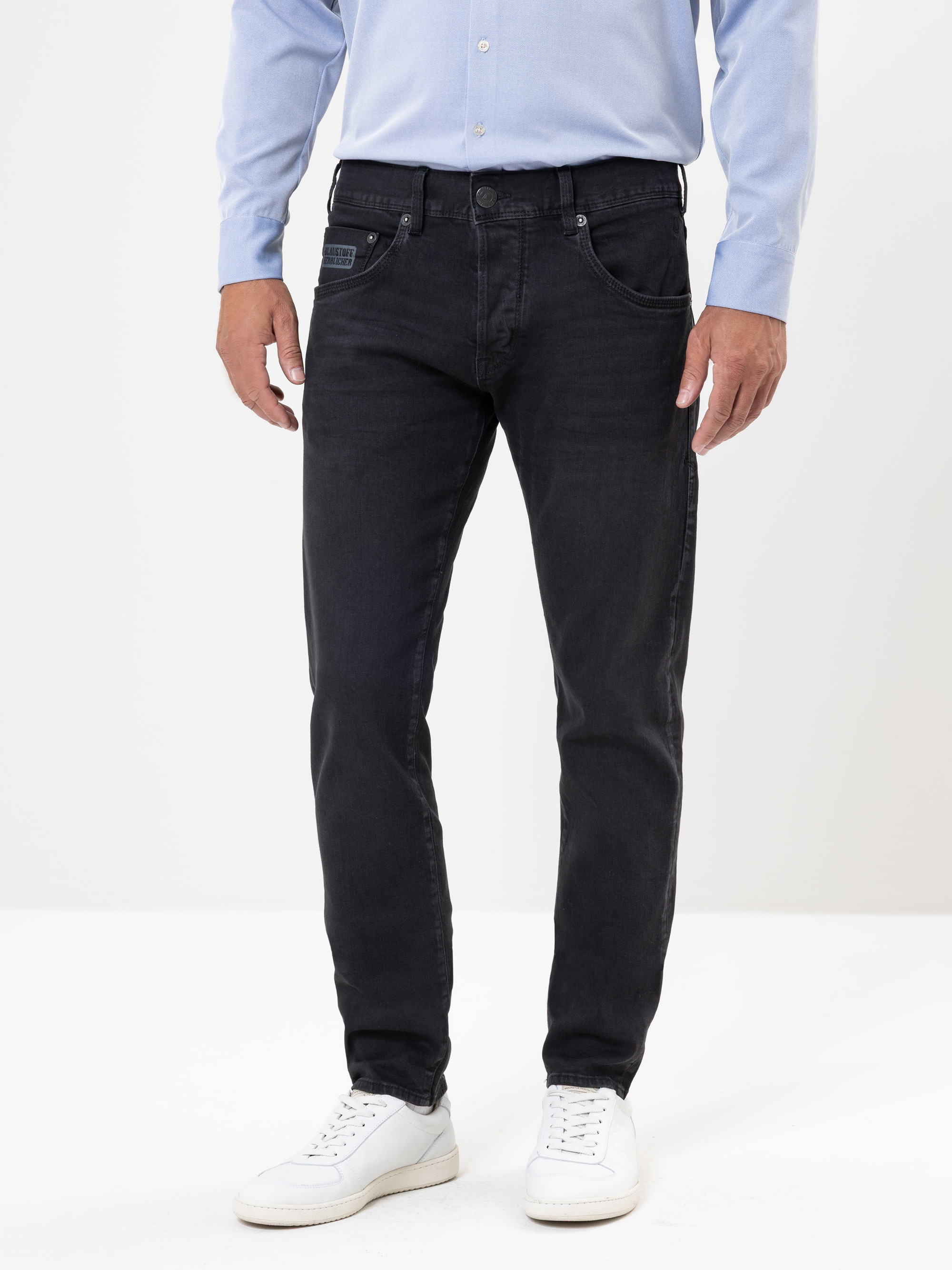 Eine Person, die ein hellblaues Button-up-Hemd, schwarze Jeans und weiße Turnschuhe trägt, steht vor einem schlichten weißen Hintergrund. Das Foto ist an den Schultern abgeschnitten und konzentriert sich auf das Outfit.