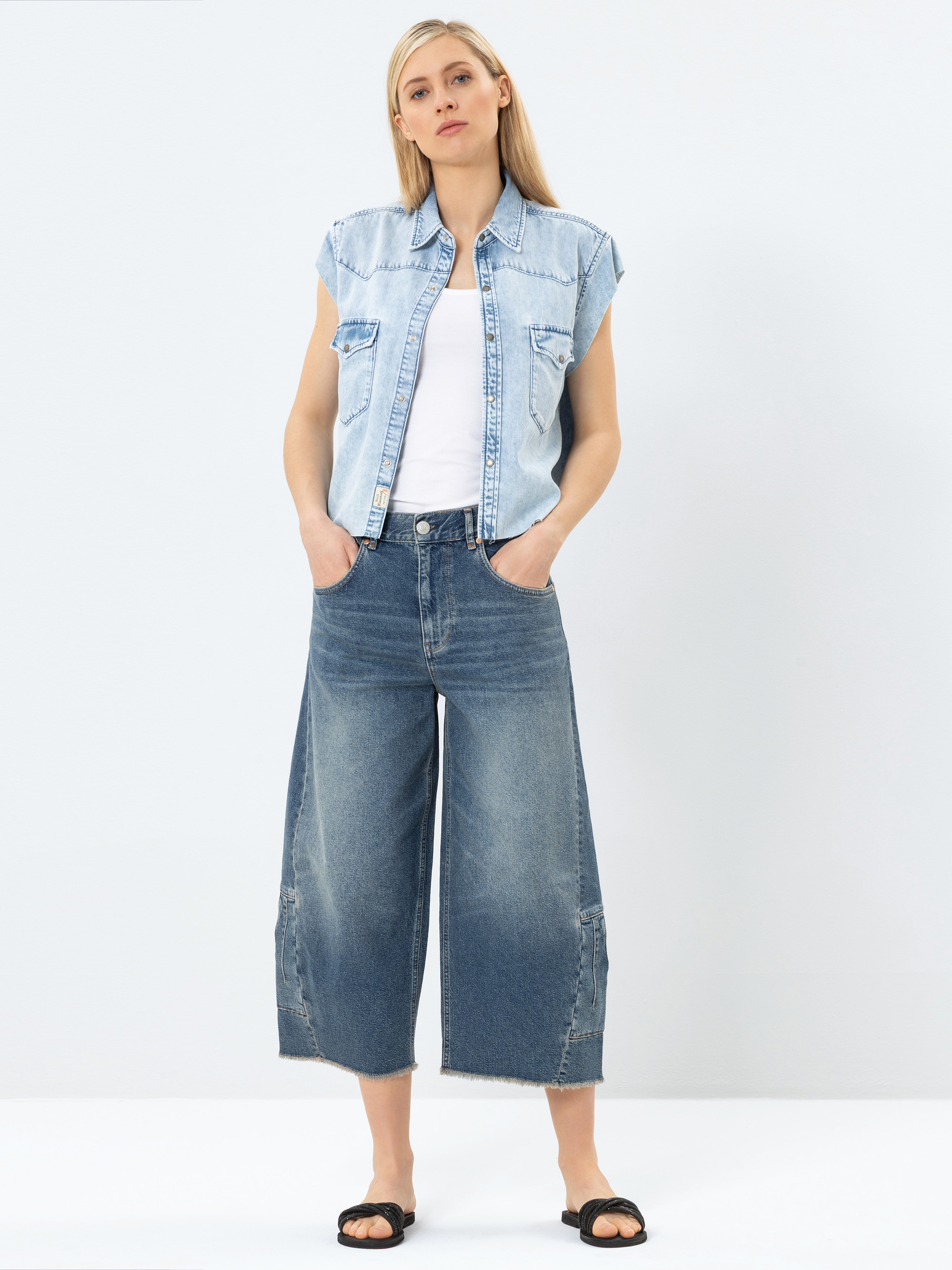 Eine Frau steht vor einem schlichten Hintergrund und trägt ein ärmelloses helles Jeanshemd über einem weißen Oberteil, weite Jeans und schwarze Sandalen. Sie hat blondes Haar und die Hände in den Taschen.