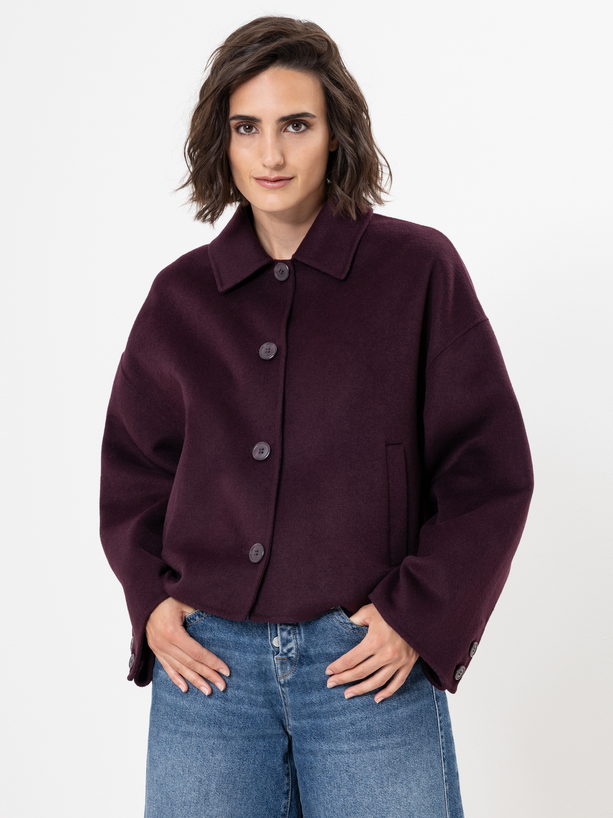 Eine Frau mit kurzen braunen Haaren, die einen dunkelvioletten Button-up-Mantel und blaue Jeans trägt, steht mit dem Gesicht zur Kamera vor einem schlichten weißen Hintergrund.