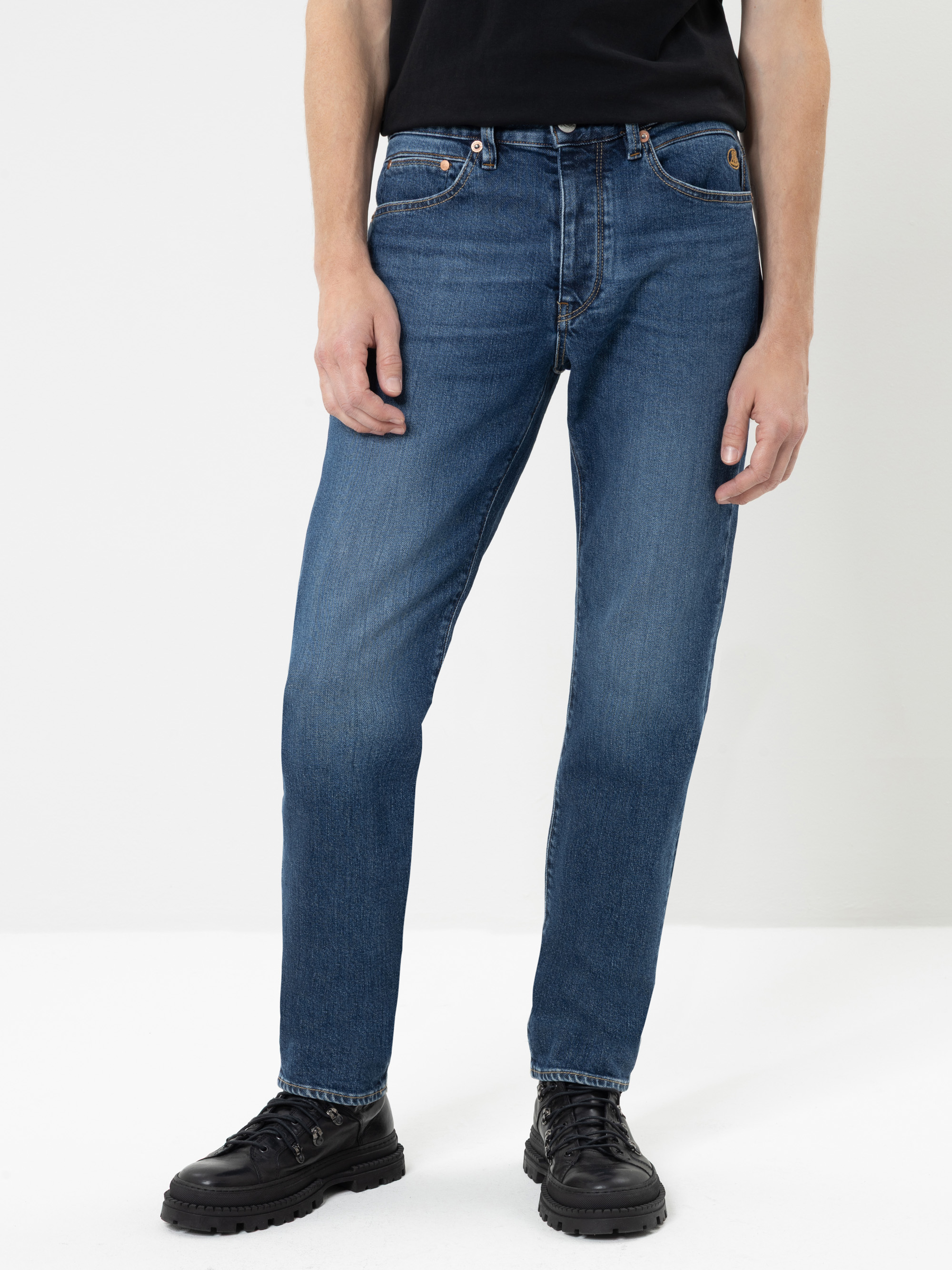 Eine Person, die eine blaue Jeans mit geradem Bein, ein schwarzes T-Shirt und schwarze Schnürschuhe trägt, steht vor einem schlichten weißen Hintergrund.