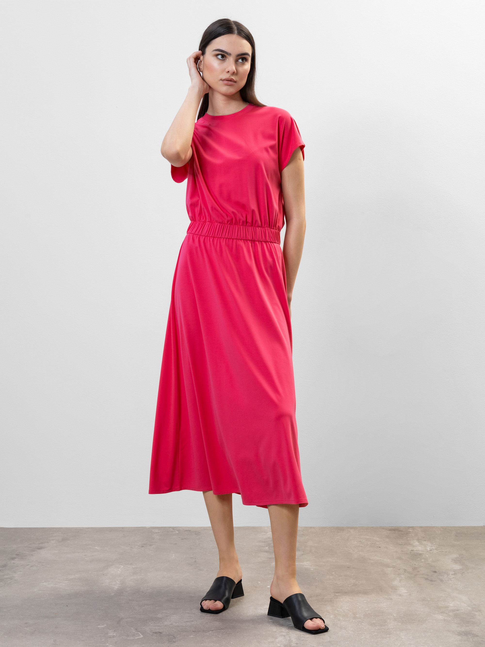 Eine Frau steht vor einem schlichten hellgrauen Hintergrund und trägt ein hellrosa, kurzärmeliges Midikleid mit elastischer Taille und schwarze Gleitsandalen. Sie berührt ihr Haar und schaut leicht zur Seite.