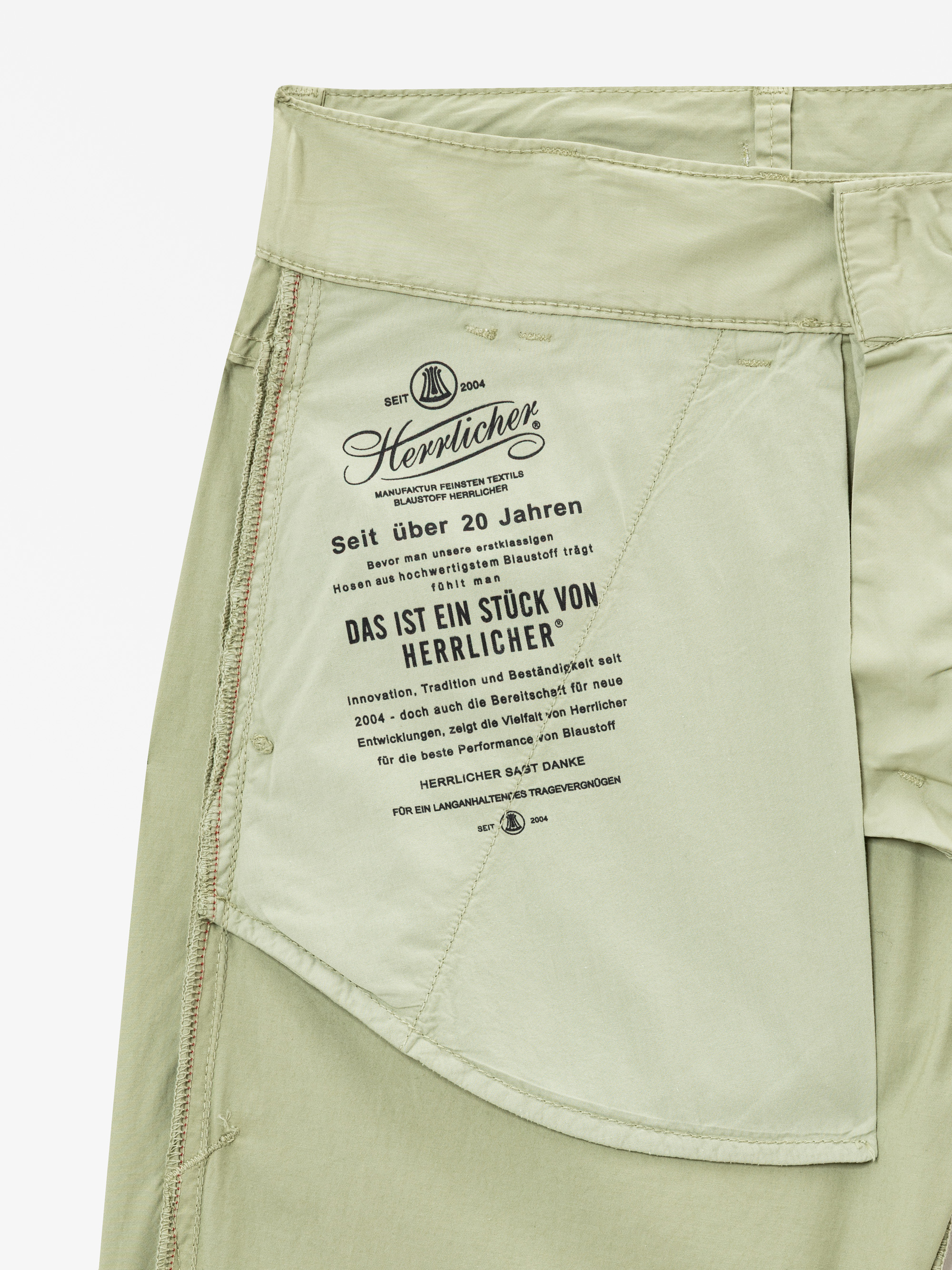 Nahaufnahme der Innentasche einer hellgrünen Hose mit einem schwarz gedruckten Etikett mit deutschem Text über die Marke Herrlicher, ihre Geschichte und Qualitätsgarantien.