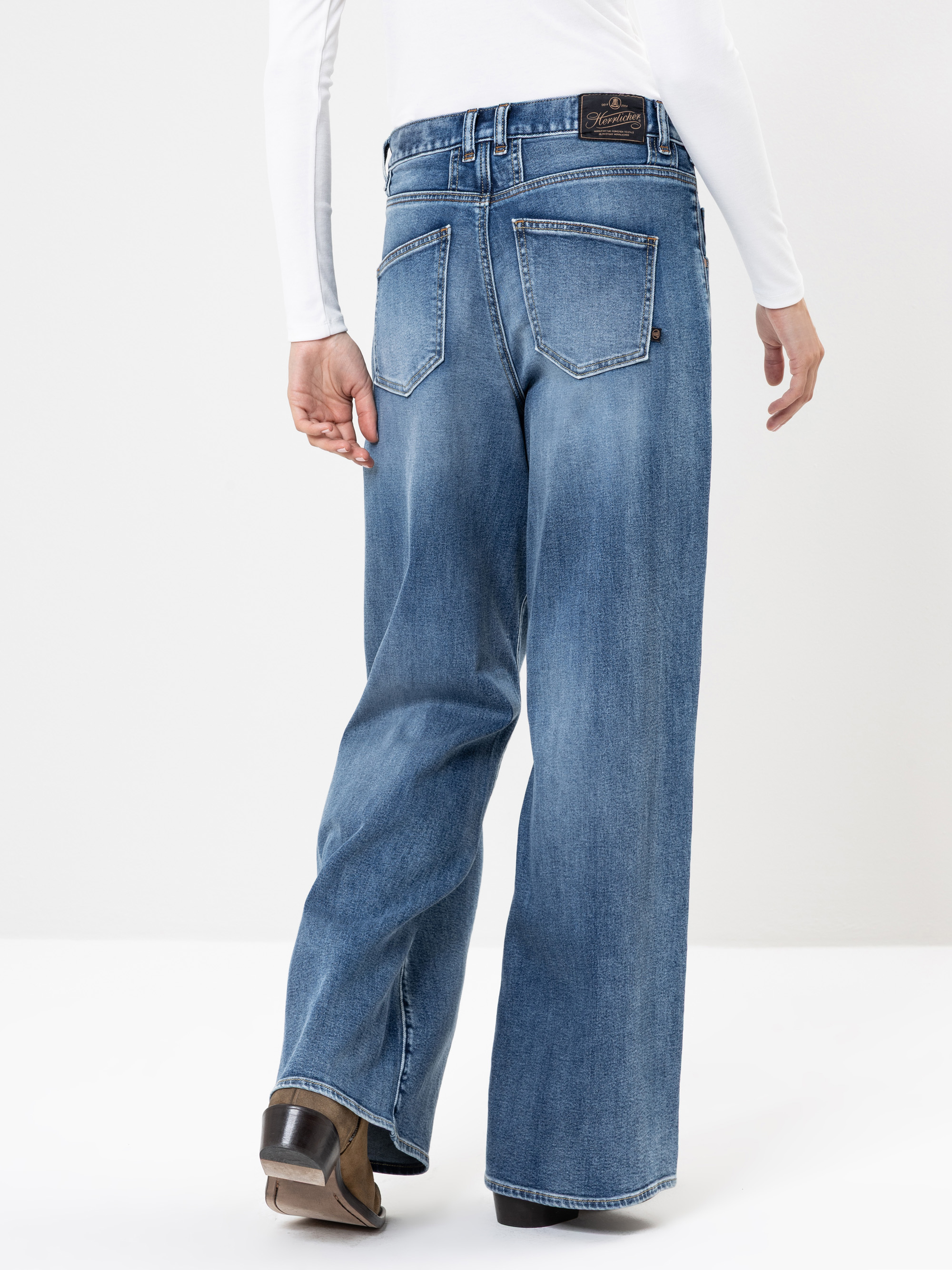 Eine Person, die eine blaue Jeans mit weitem Bein und ein weißes, langärmeliges Oberteil trägt, wird von hinten gezeigt und steht auf einem weißen Hintergrund. Die Jeans hat sichtbare Taschen und braune Schuhe sind teilweise sichtbar.