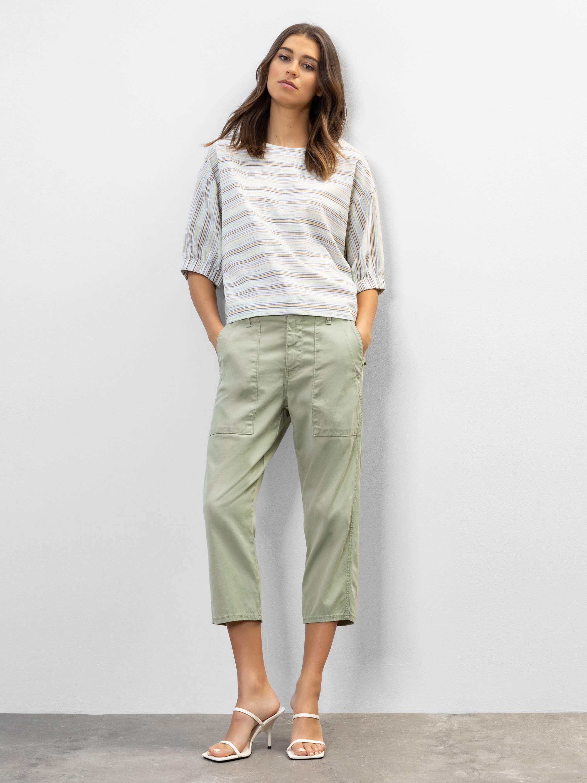 Eine Frau steht vor einer schlichten hellen Wand. Sie trägt ein lockeres gestreiftes Oberteil mit ellenbogenlangen Ärmeln, eine helle salbeigrüne Hose und weiße hochhackige Sandalen. Ihre Hände sind in den Taschen und sie schaut in die Kamera.