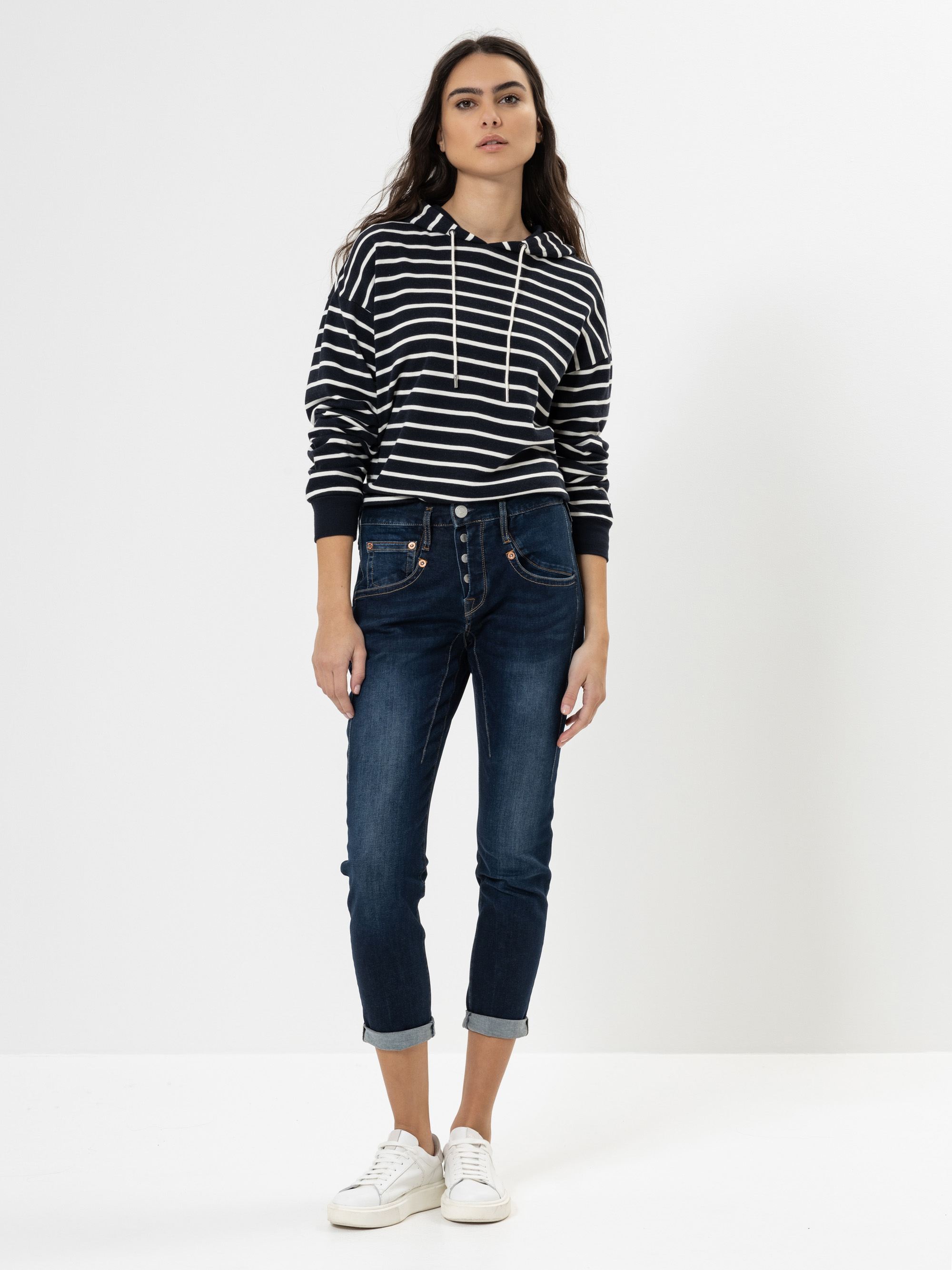 Eine Frau steht vor einem weißen Hintergrund und trägt einen schwarz-weiß gestreiften Kapuzenpullover, dunkelblaue, hochgekrempelte Jeans und weiße Turnschuhe. Sie hat langes dunkles Haar und einen neutralen Gesichtsausdruck.