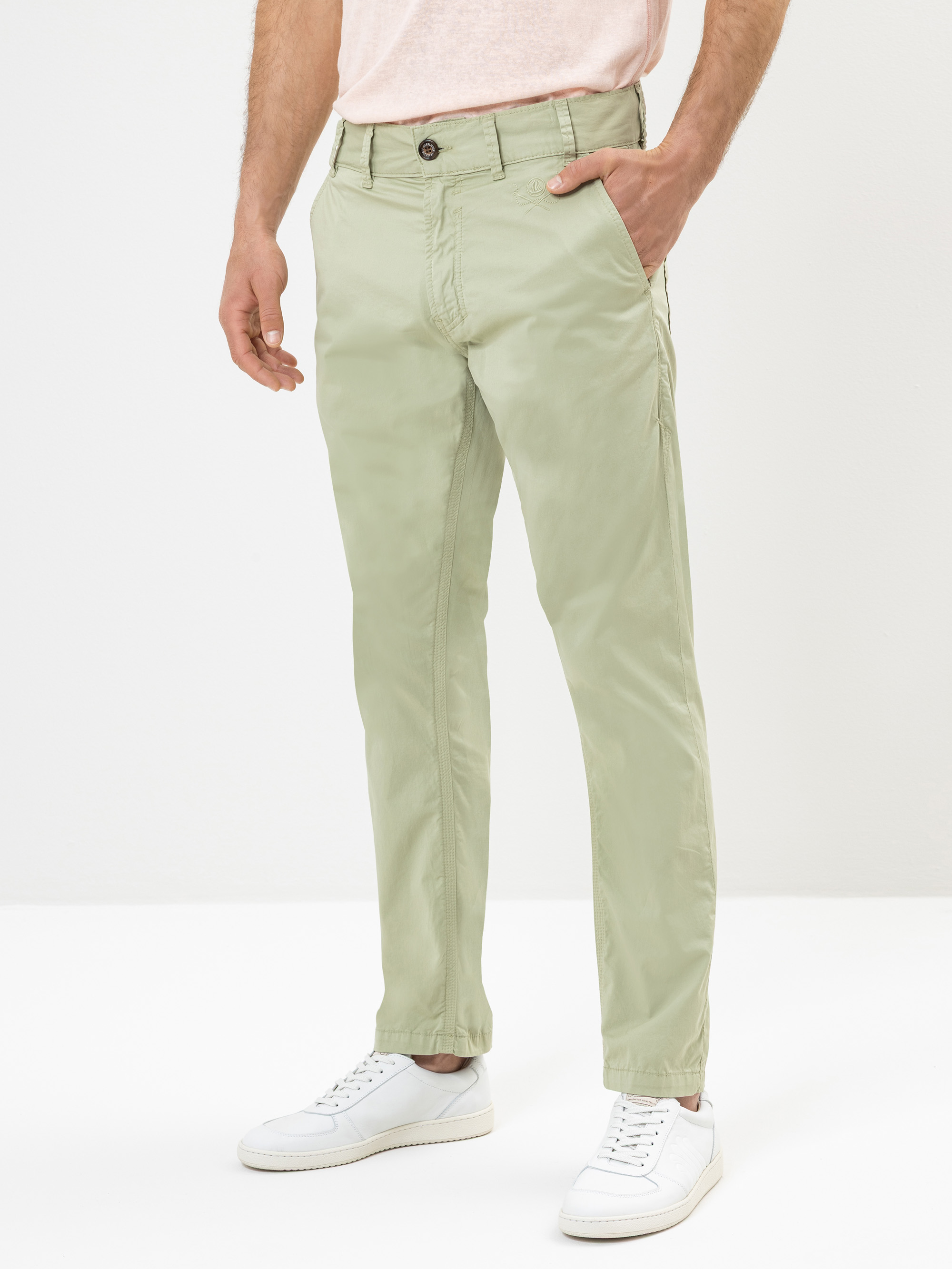Eine Person in hellgrünen Chinos, einem blassrosa T-Shirt und weißen Turnschuhen steht mit einer Hand in der Tasche vor einem schlichten weißen Hintergrund.