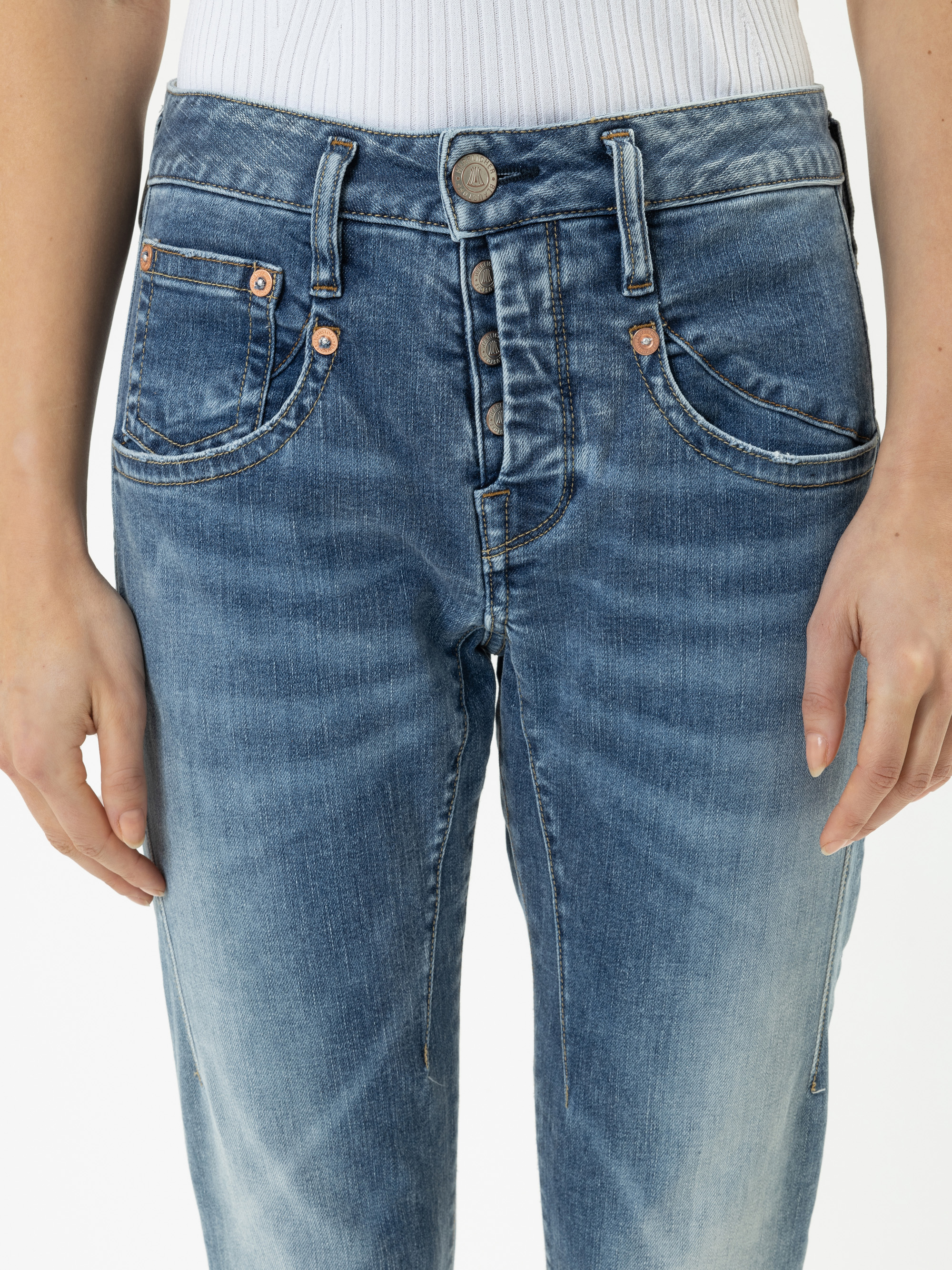 Eine Person trägt eine blaue Jeans mit Knopfleiste, die teilweise aufgeknöpft ist, und ein weißes geripptes Top. Das Bild zeigt die Taille und die Oberschenkel, die Hände liegen entspannt an den Seiten.