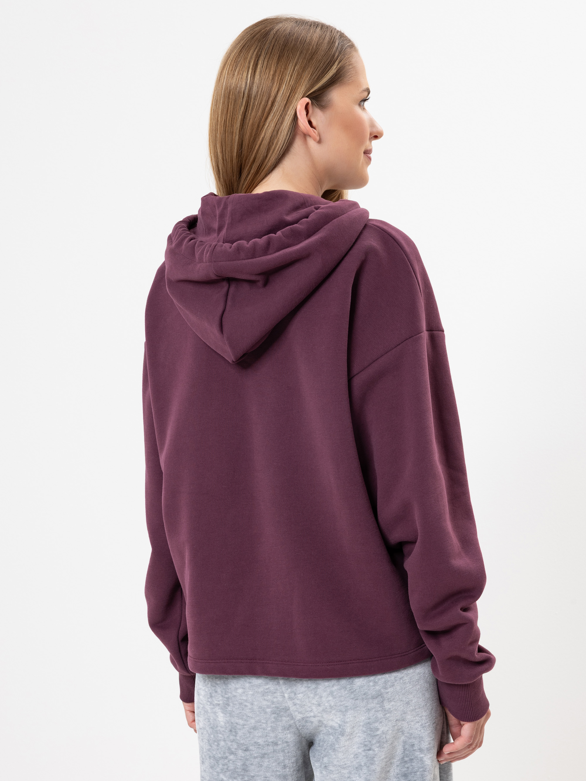 Eine Frau mit glattem, hellbraunem Haar wird von hinten gezeigt. Sie trägt einen locker sitzenden, dunkelvioletten Kapuzenpullover und eine hellgraue Hose und steht vor einem schlichten weißen Hintergrund.