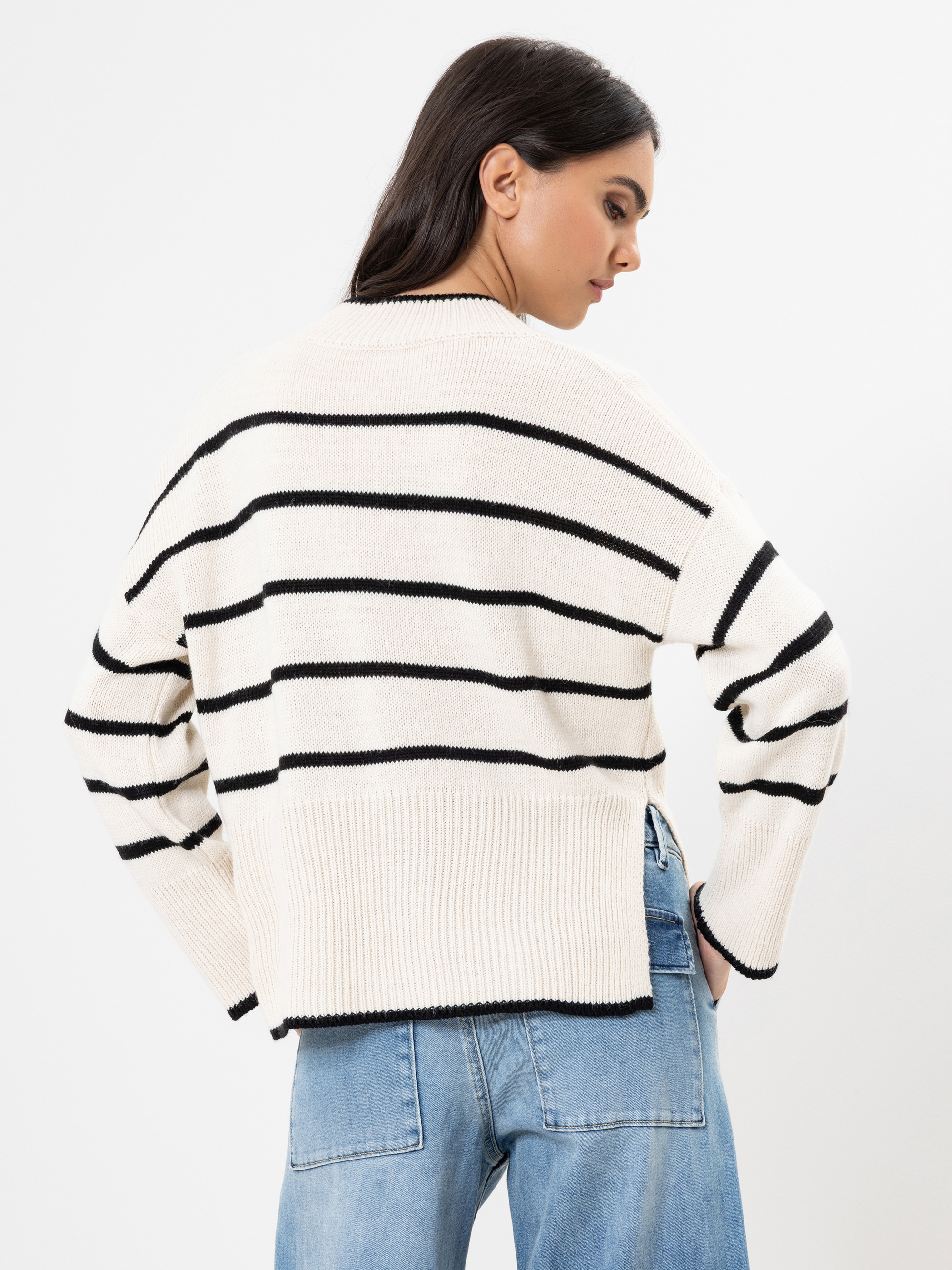 Eine Frau mit langen dunklen Haaren steht mit dem Gesicht zur Wand. Sie trägt einen cremefarbenen Pullover mit schwarzen Querstreifen und hellblaue Jeans vor einem schlichten weißen Hintergrund.
