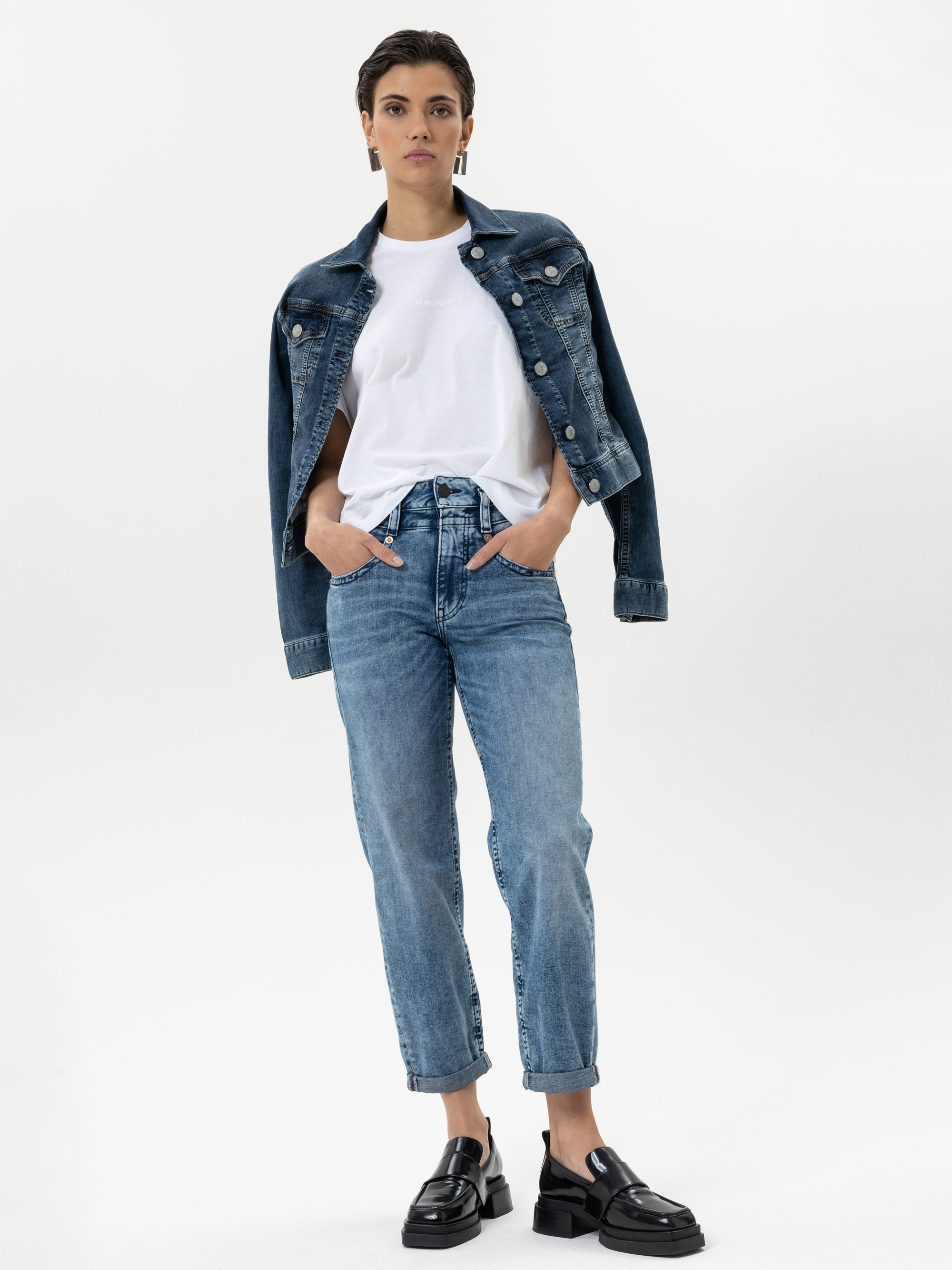 Eine Frau steht mit den Händen in den Taschen, trägt ein weißes T-Shirt, eine blaue Jeansjacke, die sie über die Schultern gelegt hat, blaue Jeans mit Bündchen und schwarze Slipper vor einem schlichten weißen Hintergrund.