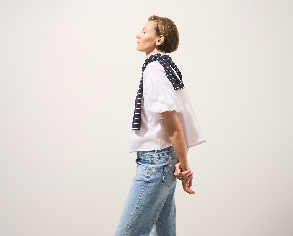 Eine Person mit kurzen Haaren steht im Profil vor einem schlichten Hintergrund, die Augen geschlossen, mit weißer Bluse, hellblauer Jeans und einem marineblau gestreiften Pullover, der über die Schultern gelegt ist, die Hände hinter dem Rücken verschr