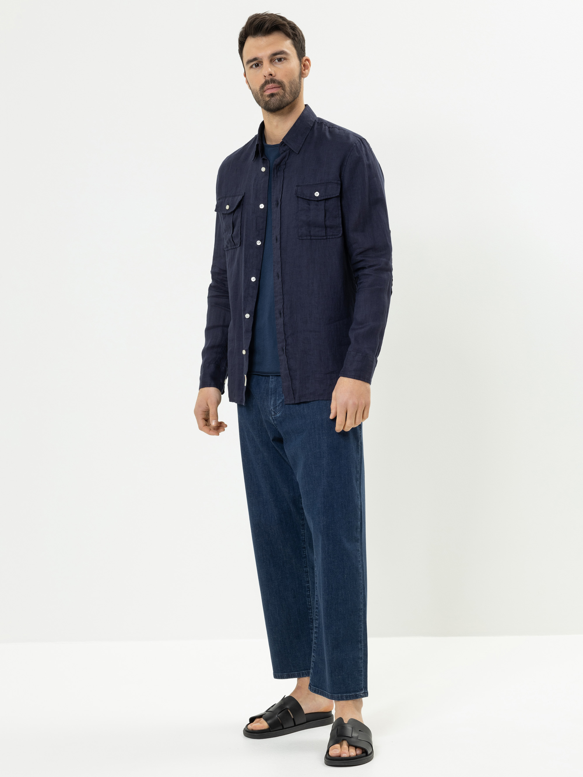 Ein Mann steht vor einem weißen Hintergrund und trägt ein marineblaues Button-up-Hemd über einem T-Shirt, locker sitzende Bluejeans und schwarze Sandalen. Er hat kurzes dunkles Haar, einen Bart und einen neutralen Gesichtsausdruck.