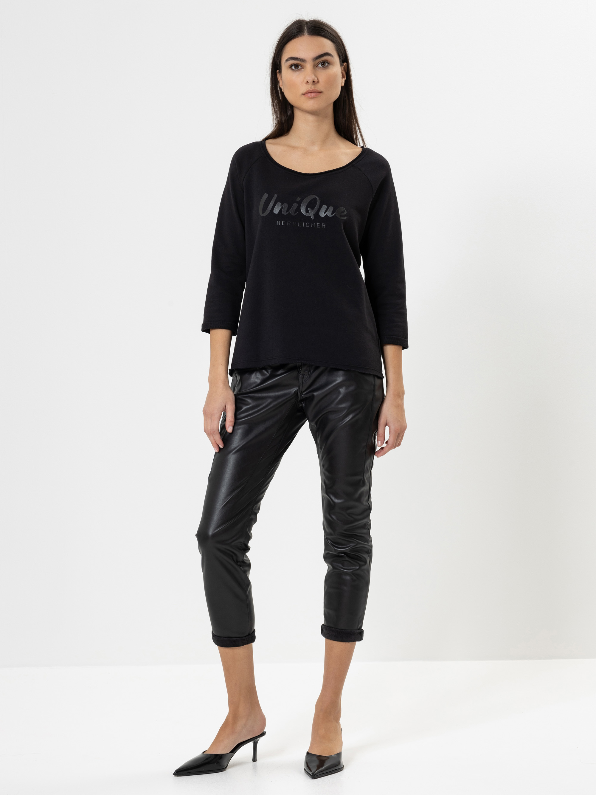 Eine Frau steht vor einem weißen Hintergrund und trägt ein schwarzes langärmeliges Oberteil mit der Aufschrift Unique, eine schwarze Lederhose und schwarze spitze Absätze. Sie hat langes dunkles Haar und einen neutralen Gesichtsausdruck.