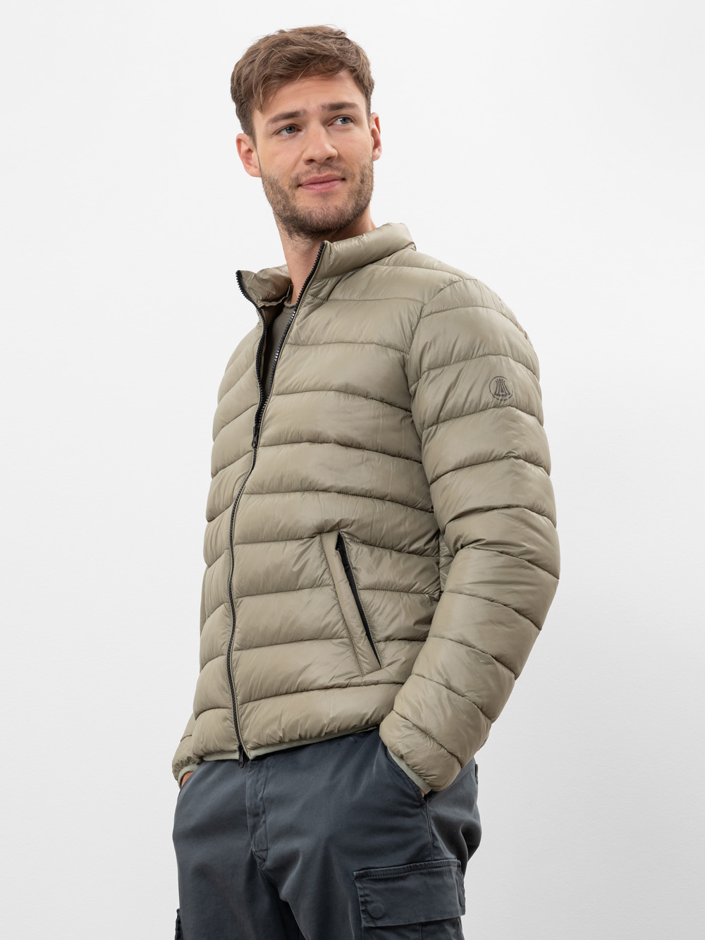 Ein Mann, der eine helle olivgrüne Pufferjacke mit Reißverschluss und eine dunkelgraue Cargohose trägt, steht mit den Händen in den Taschen und schaut leicht zur Seite. Der Hintergrund ist einfarbig weiß.