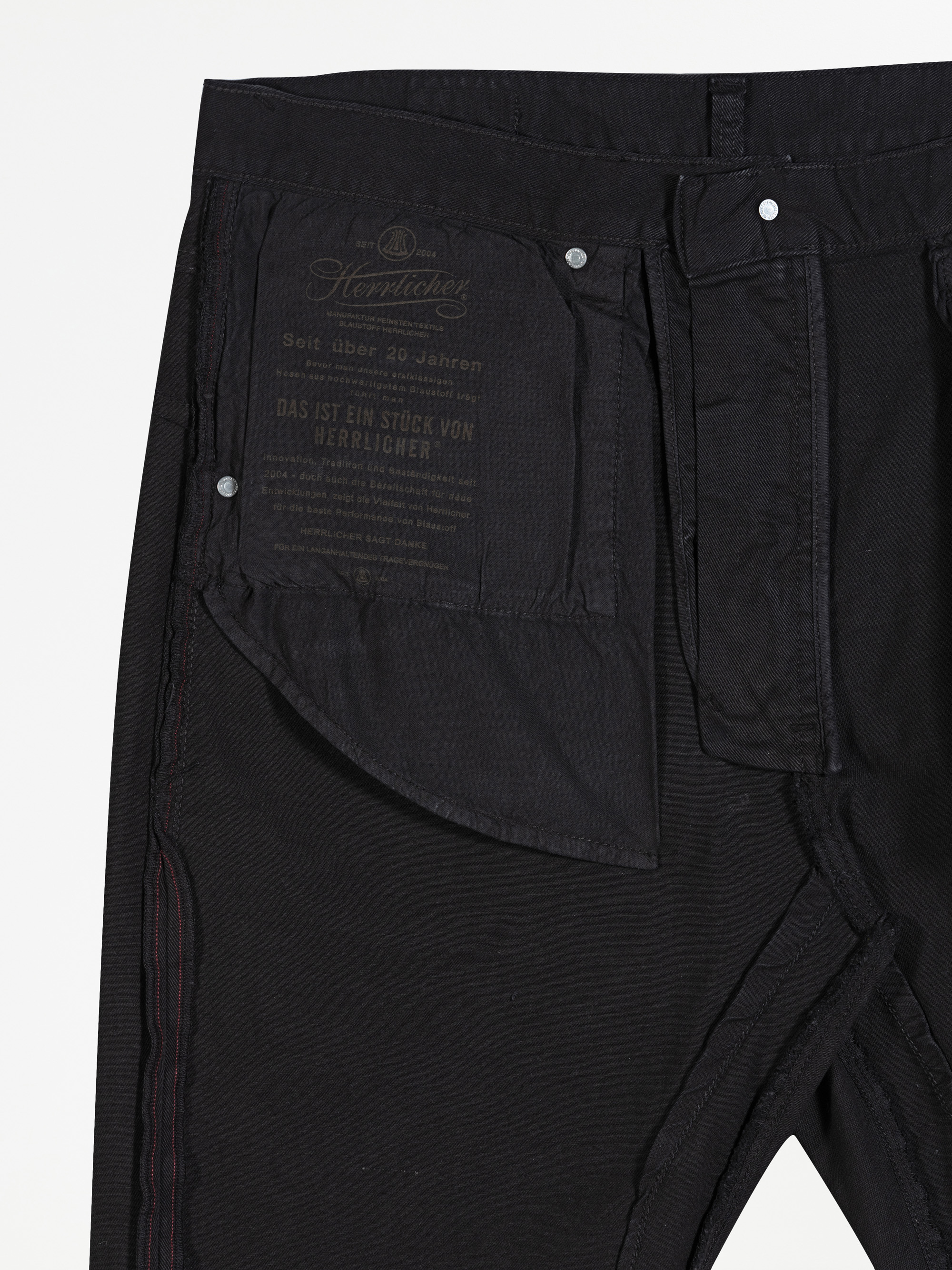 Eine Nahaufnahme des Innenbundes einer schwarzen Jeans, die einen Stoffaufnäher mit deutschem Text und Markenzeichen, silberne Nieten und detaillierte Nähte zeigt.