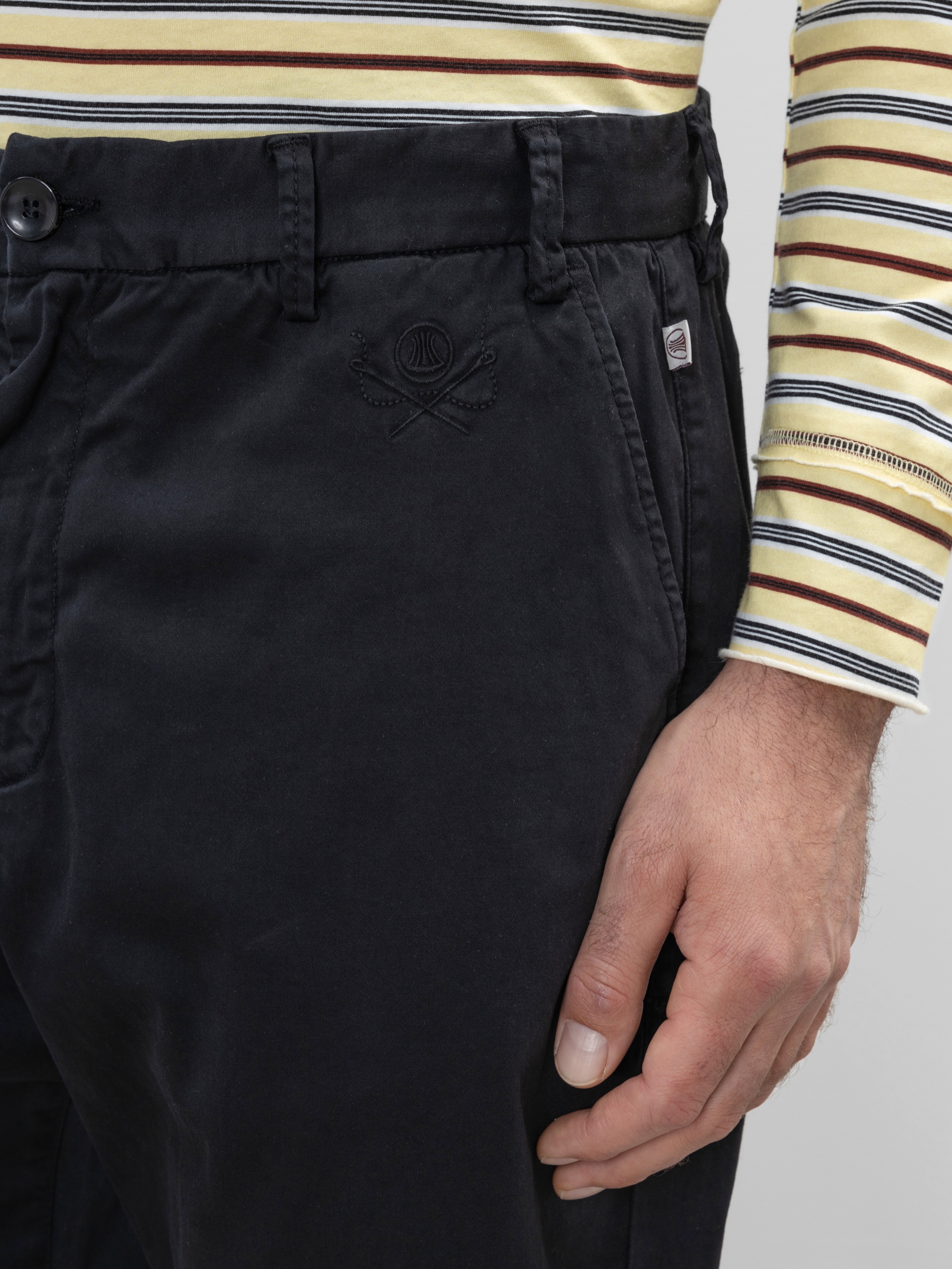 Eine Person, die eine dunkle marineblaue Hose mit einem aufgestickten Logo in der Nähe der Tasche und ein langärmeliges gelb, weiß und schwarz gestreiftes Hemd trägt. Ihre rechte Hand liegt entspannt an ihrer Seite.