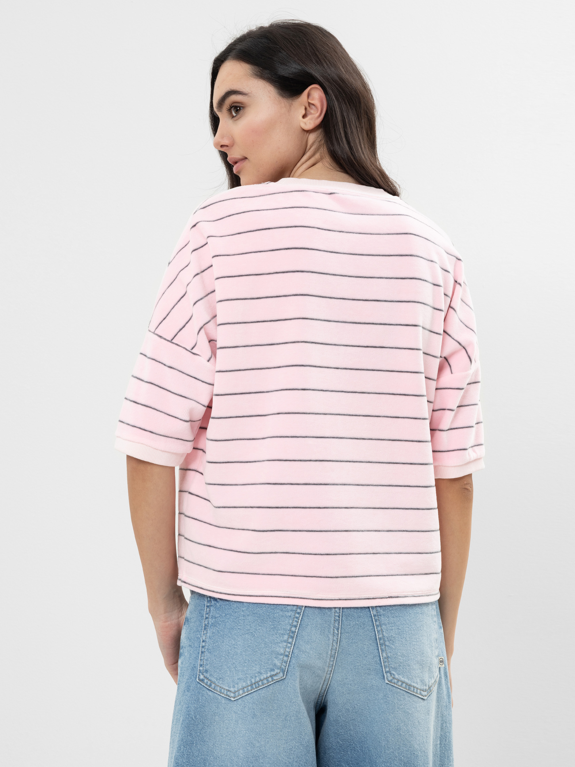 Frau mit langen dunklen Haaren trägt ein rosa-weiß gestreiftes Oberteil mit ellbogenlangen Ärmeln und hellblaue Jeans. Sie steht mit dem Rücken zur Kamera vor einem schlichten Hintergrund.