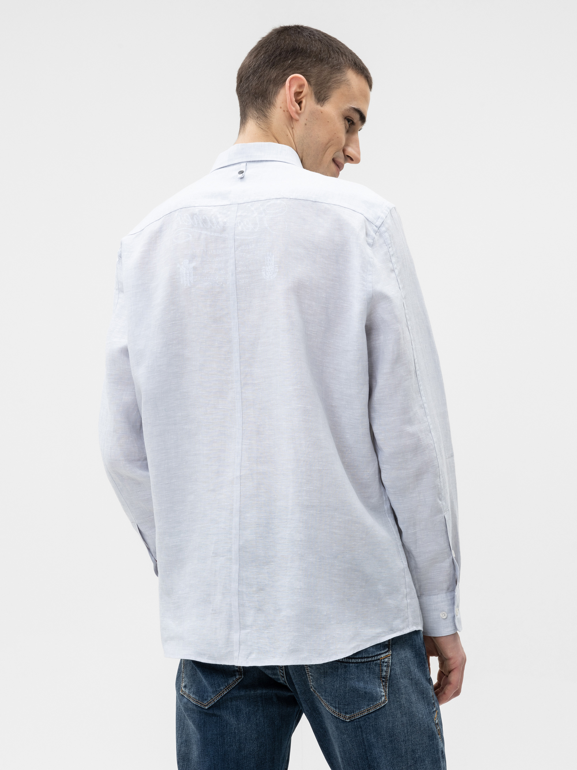 Ein Mann mit kurzen braunen Haaren trägt ein helles, langärmeliges Button-up-Hemd und blaue Jeans. Er ist leicht zur Seite gedreht und blickt über seine Schulter auf einen schlichten weißen Hintergrund.