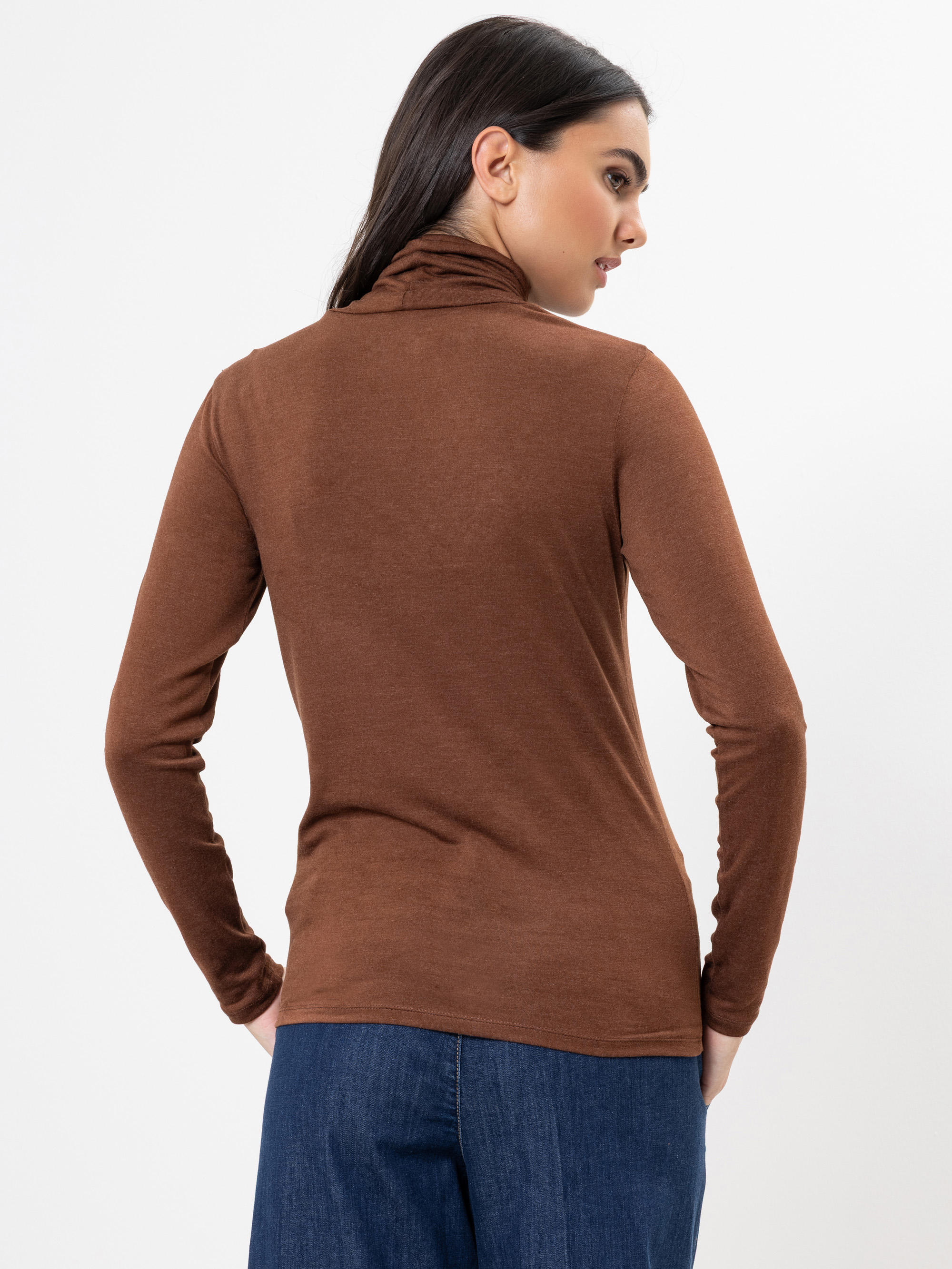 Eine Frau mit glattem dunklem Haar trägt ein braunes langärmeliges Rollkragenoberteil und blaue Jeans. Sie blickt von der Kamera weg nach links, vor einem schlichten weißen Hintergrund.