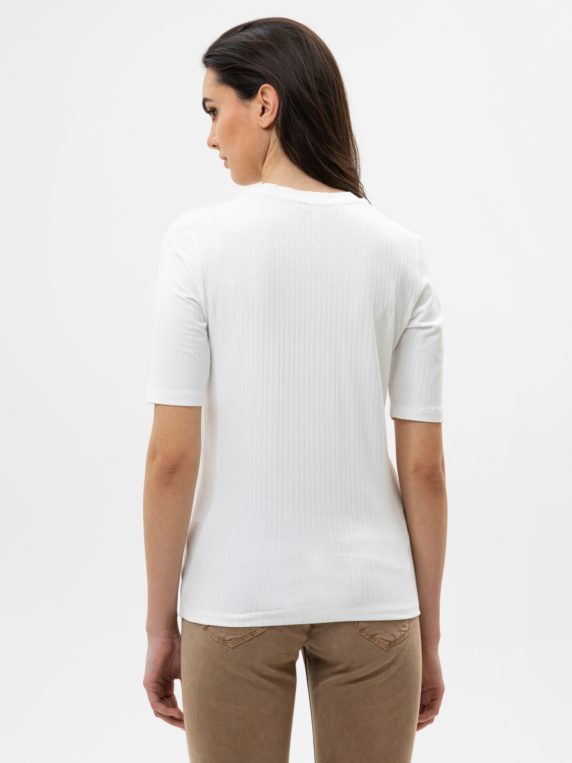 Frau mit langen dunklen Haaren, von hinten gesehen, trägt ein weißes kurzärmeliges geripptes Oberteil und eine beigefarbene Hose und steht vor einem schlichten weißen Hintergrund.