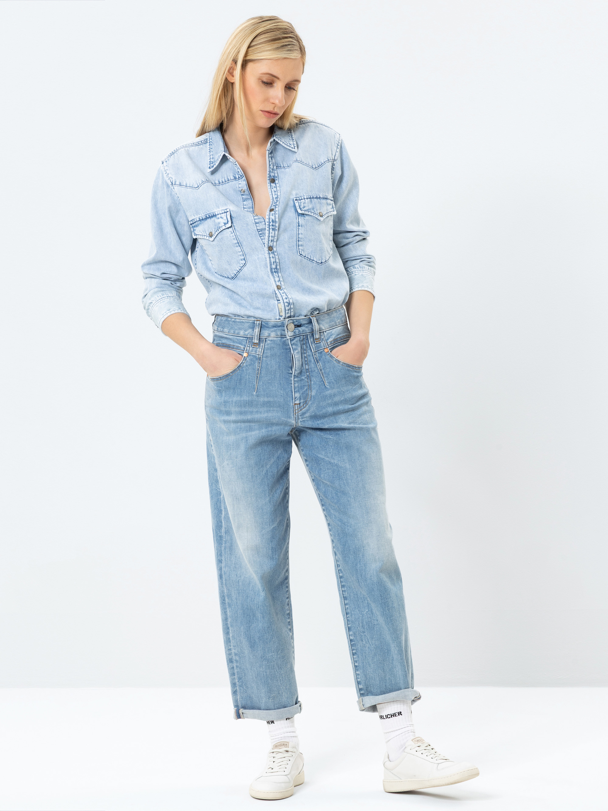 Eine Frau steht mit den Händen in den Taschen und trägt ein hellblaues Jeanshemd, das in eine hoch taillierte, locker sitzende Bluejeans gesteckt wird, weiße Socken und weiße Turnschuhe vor einem schlichten weißen Hintergrund.