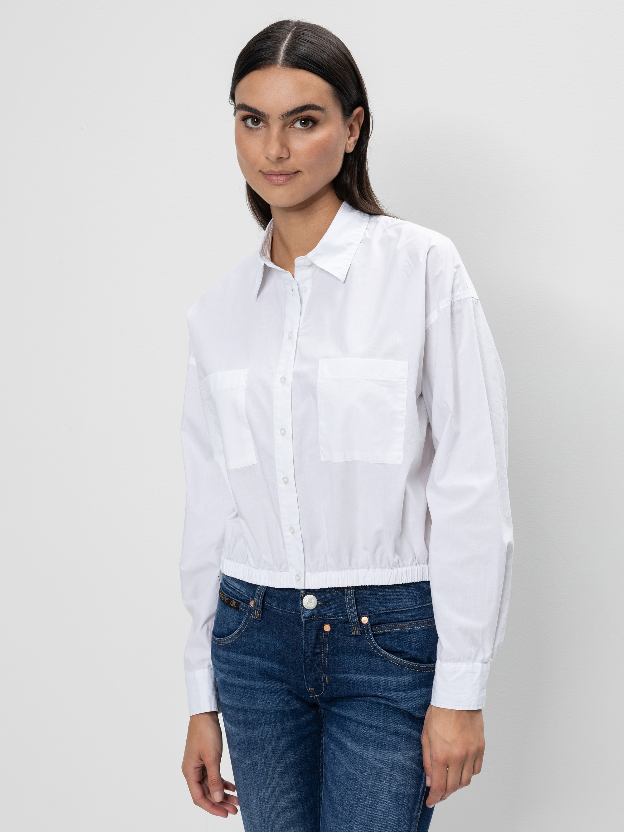 Eine Frau mit glattem dunklem Haar trägt ein weißes Button-up-Hemd mit Brusttaschen und blaue Jeans. Sie steht vor einem schlichten weißen Hintergrund und blickt mit einem leichten Lächeln nach vorne.