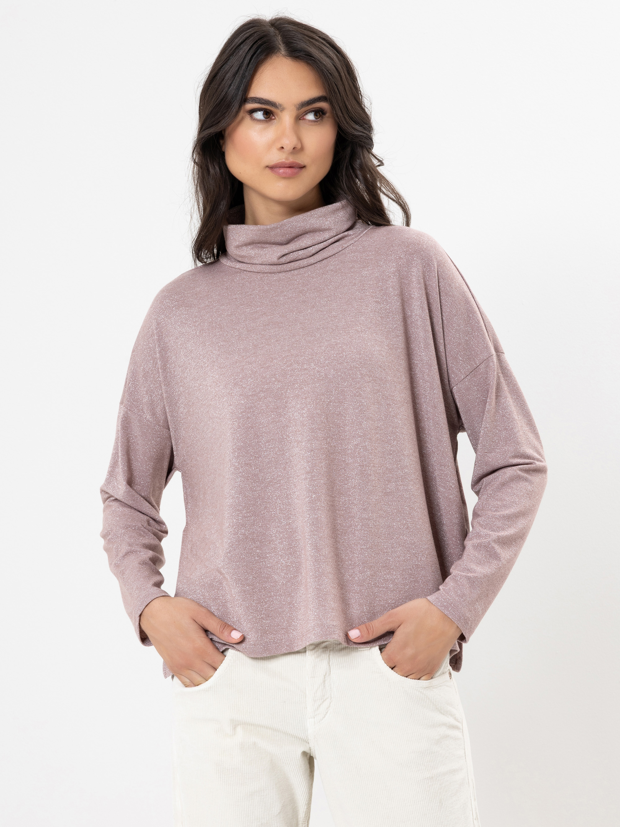 Eine Frau mit langen dunklen Haaren trägt einen helllilafarbenen Rollkragenpullover und eine helle Hose. Sie steht vor einem schlichten weißen Hintergrund, hat die Hände in den Taschen und schaut leicht zur Seite.