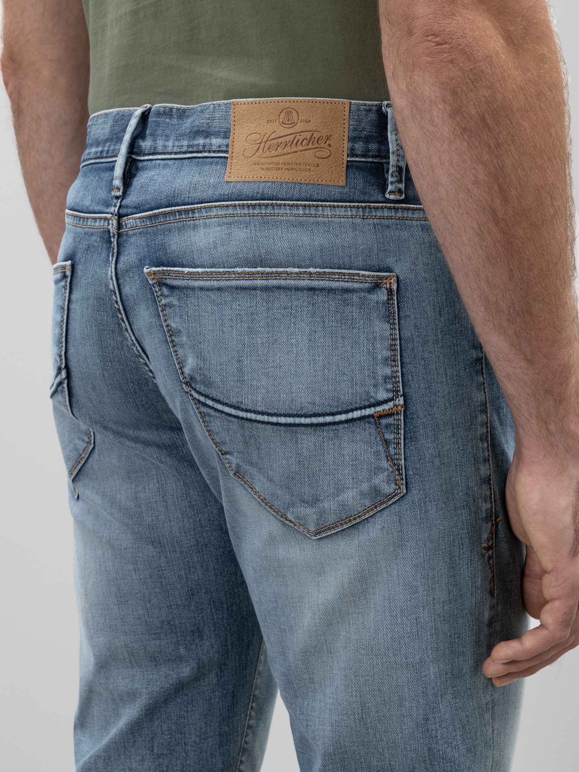 Nahaufnahme einer Person, die eine blaue Jeans trägt, wobei die Gesäßtasche und ein braunes Markenetikett aus Leder am Hosenbund zu sehen sind. Die Person steht seitlich, ihr Arm ist sichtbar.