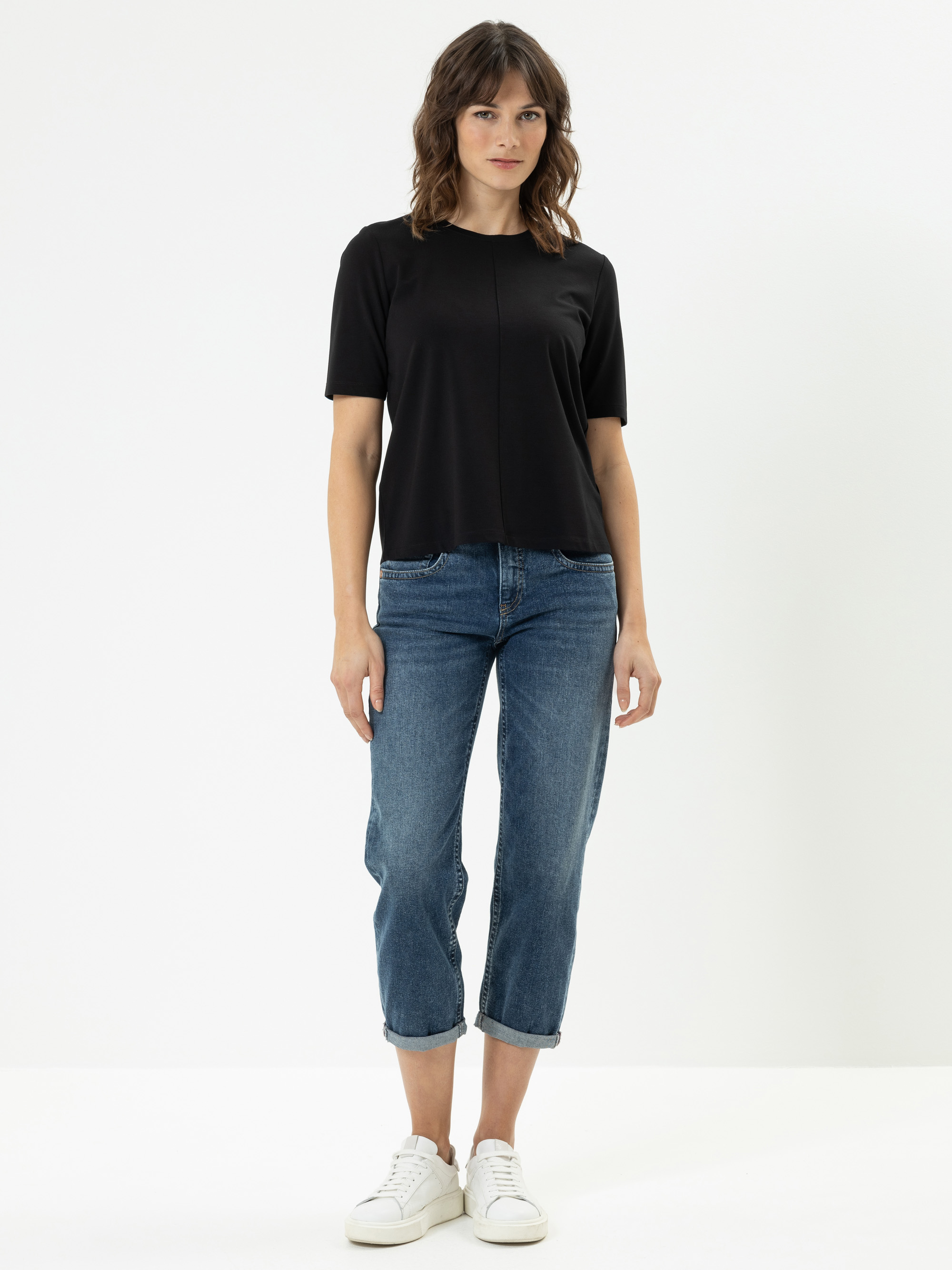 Eine Frau steht vor einem schlichten weißen Hintergrund und trägt ein schwarzes, kurzärmeliges Oberteil, eine blaue, kurze Jeans und weiße Turnschuhe. Sie hat gewelltes braunes Haar und einen neutralen Gesichtsausdruck.