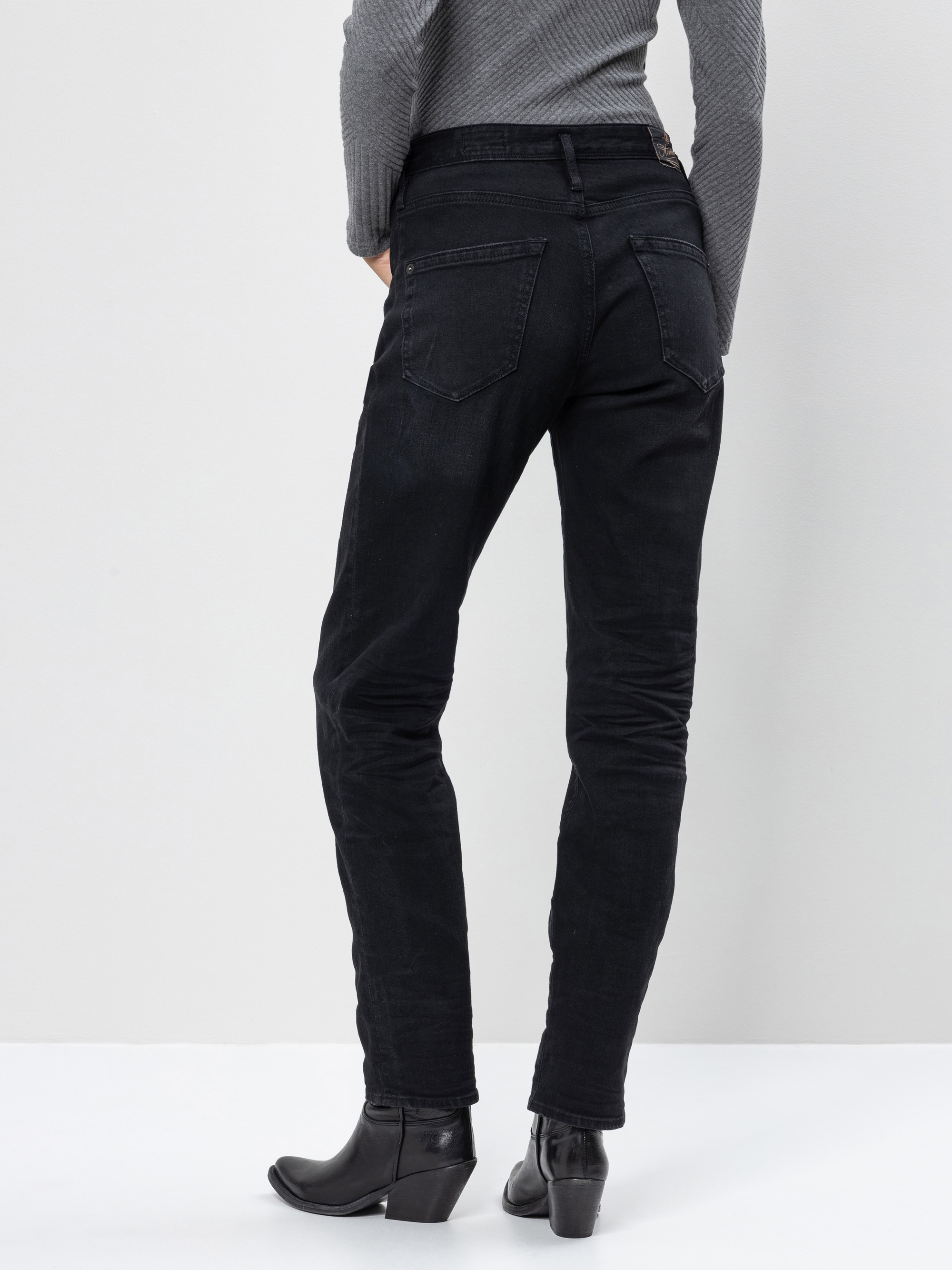 Eine Person, die schwarze Jeans, ein graues geripptes langärmeliges Oberteil und schwarze hochhackige Stiefel trägt, steht mit dem Gesicht zur Seite vor einem schlichten hellen Hintergrund.