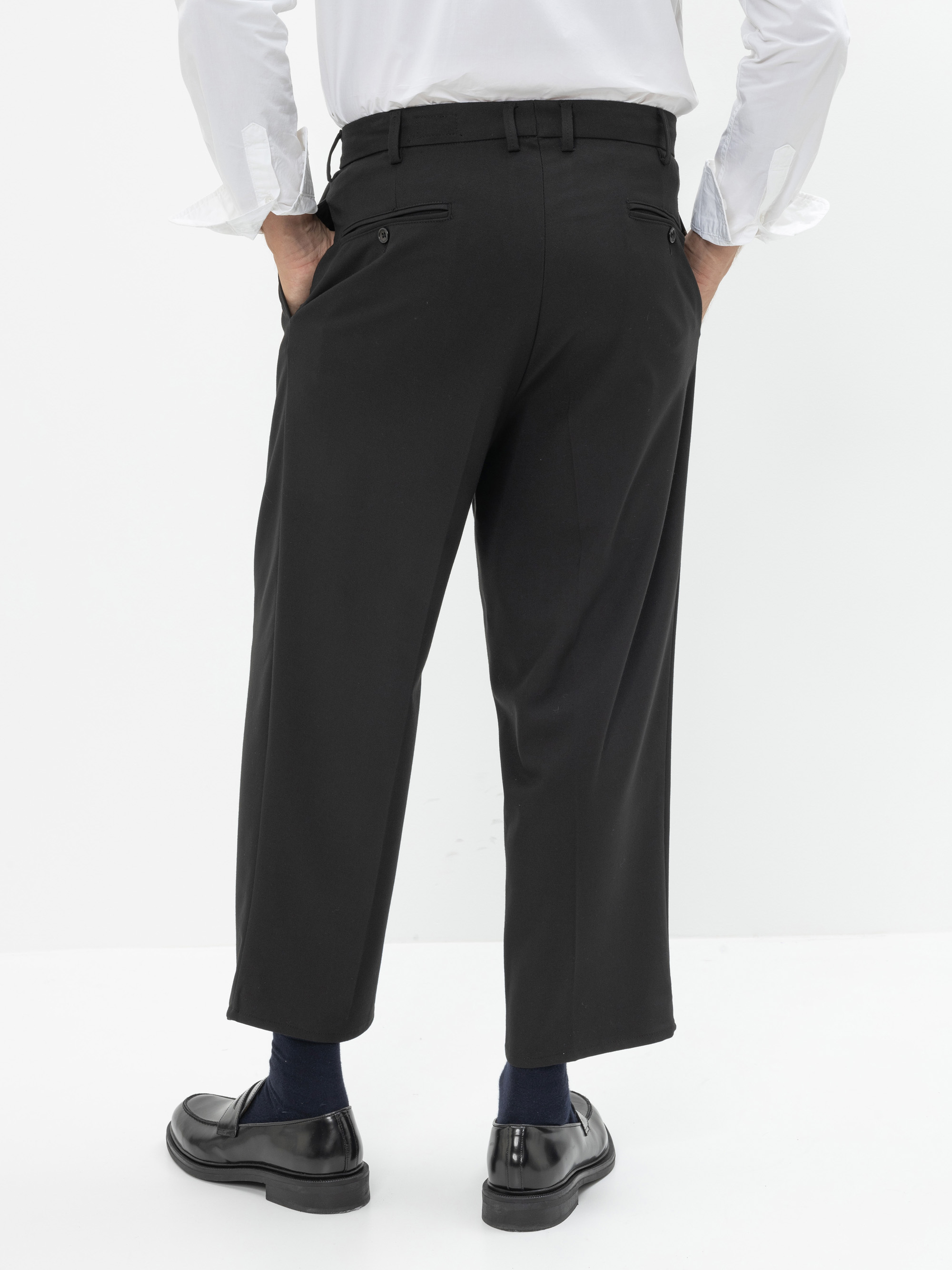 Eine Person steht mit dem Gesicht zur Wand und trägt ein weißes Hemd, eine schwarze Hose, dunkle Socken und schwarze Schuhe. Ihre Hände stecken in den Taschen vor einem schlichten weißen Hintergrund.