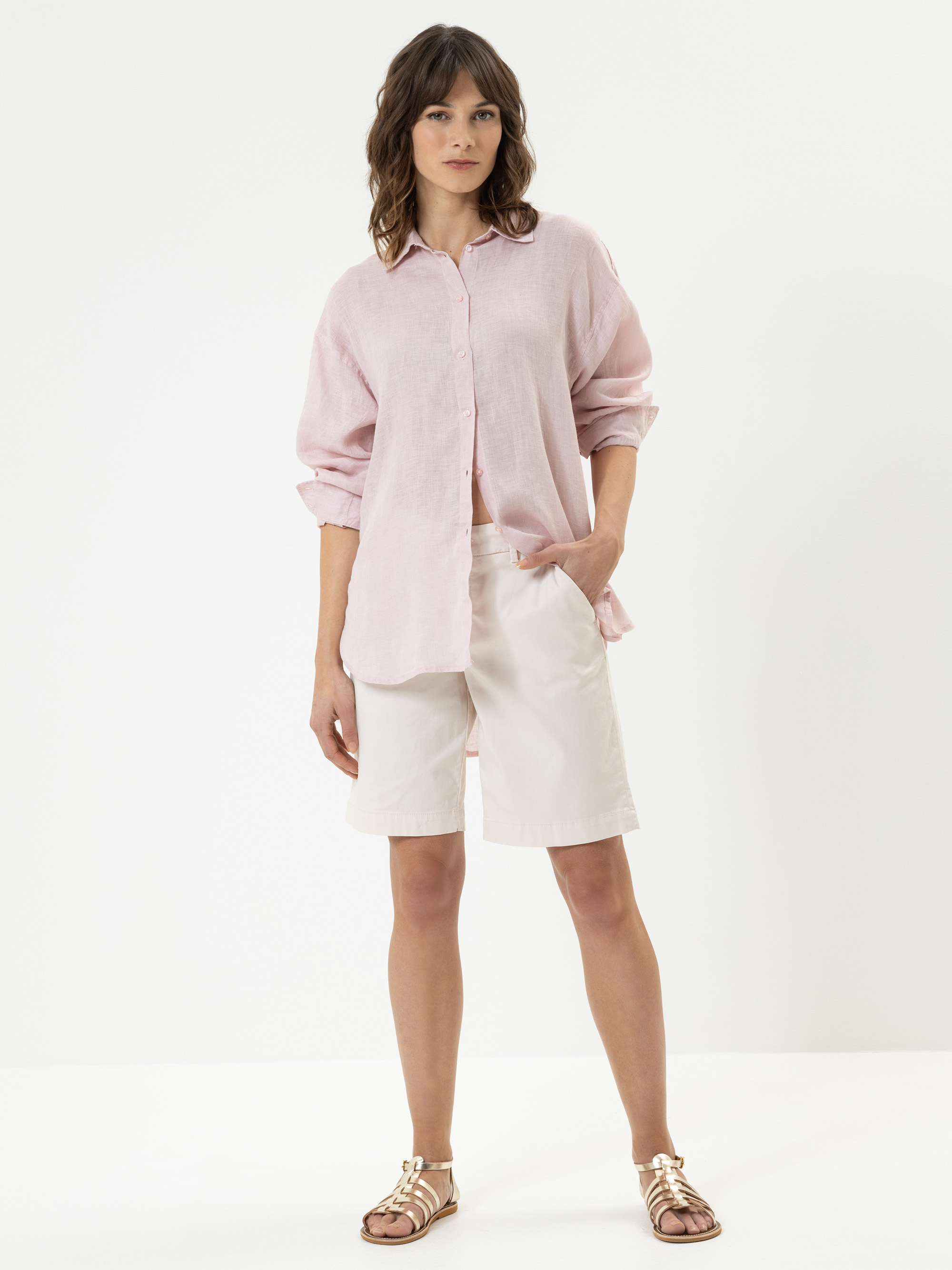 Eine Frau, die ein locker sitzendes hellrosa Button-up-Hemd, hellbeige knielange Shorts und goldene Riemchensandalen trägt, steht vor einem schlichten weißen Hintergrund.