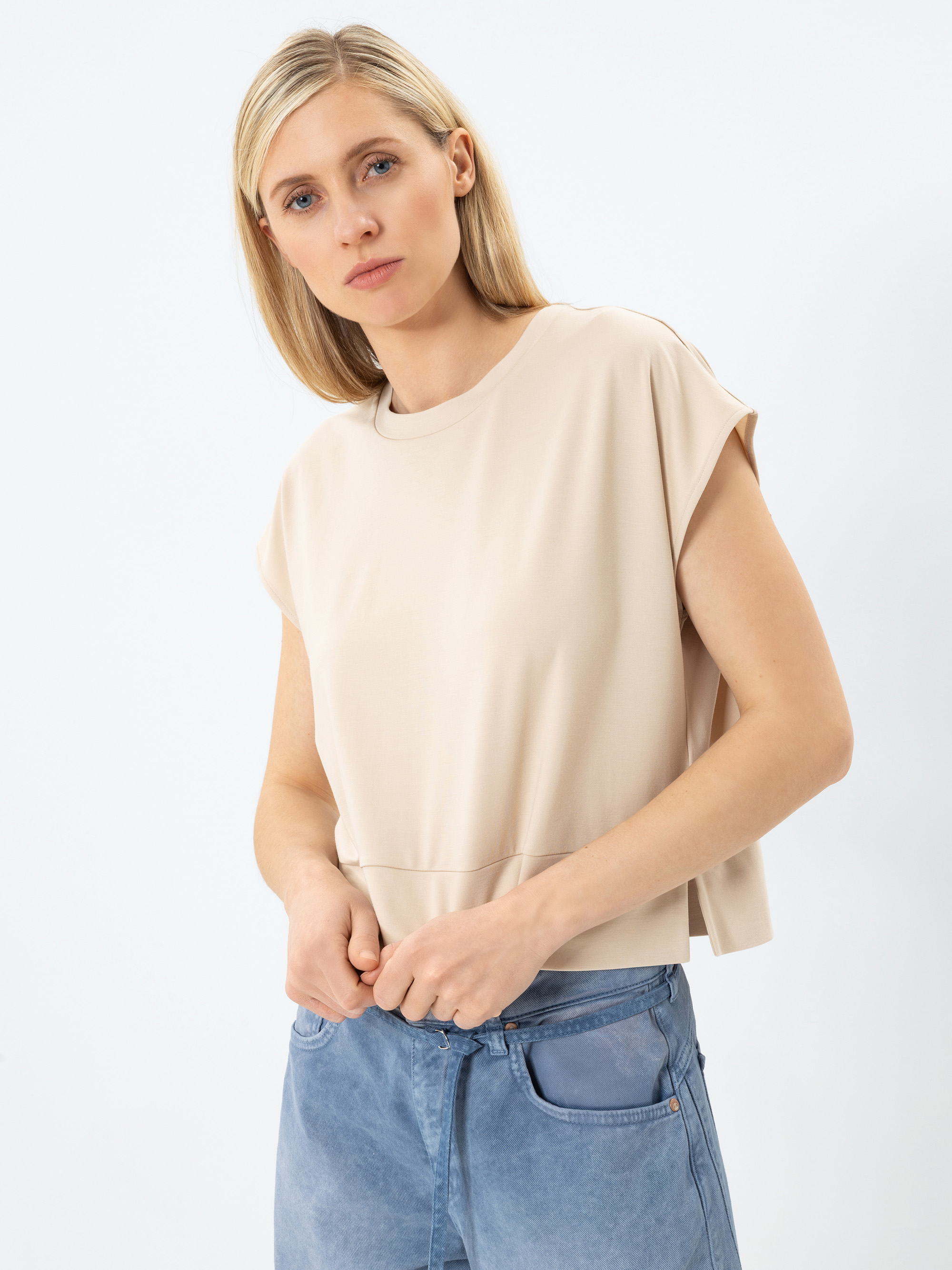 Eine Frau mit glattem blondem Haar trägt ein beigefarbenes kurzärmeliges Oberteil und hellblaue Jeans und steht vor einem schlichten weißen Hintergrund. Sie blickt leicht zur Seite mit einem neutralen Ausdruck und hält ihre Jeans in der Taille.