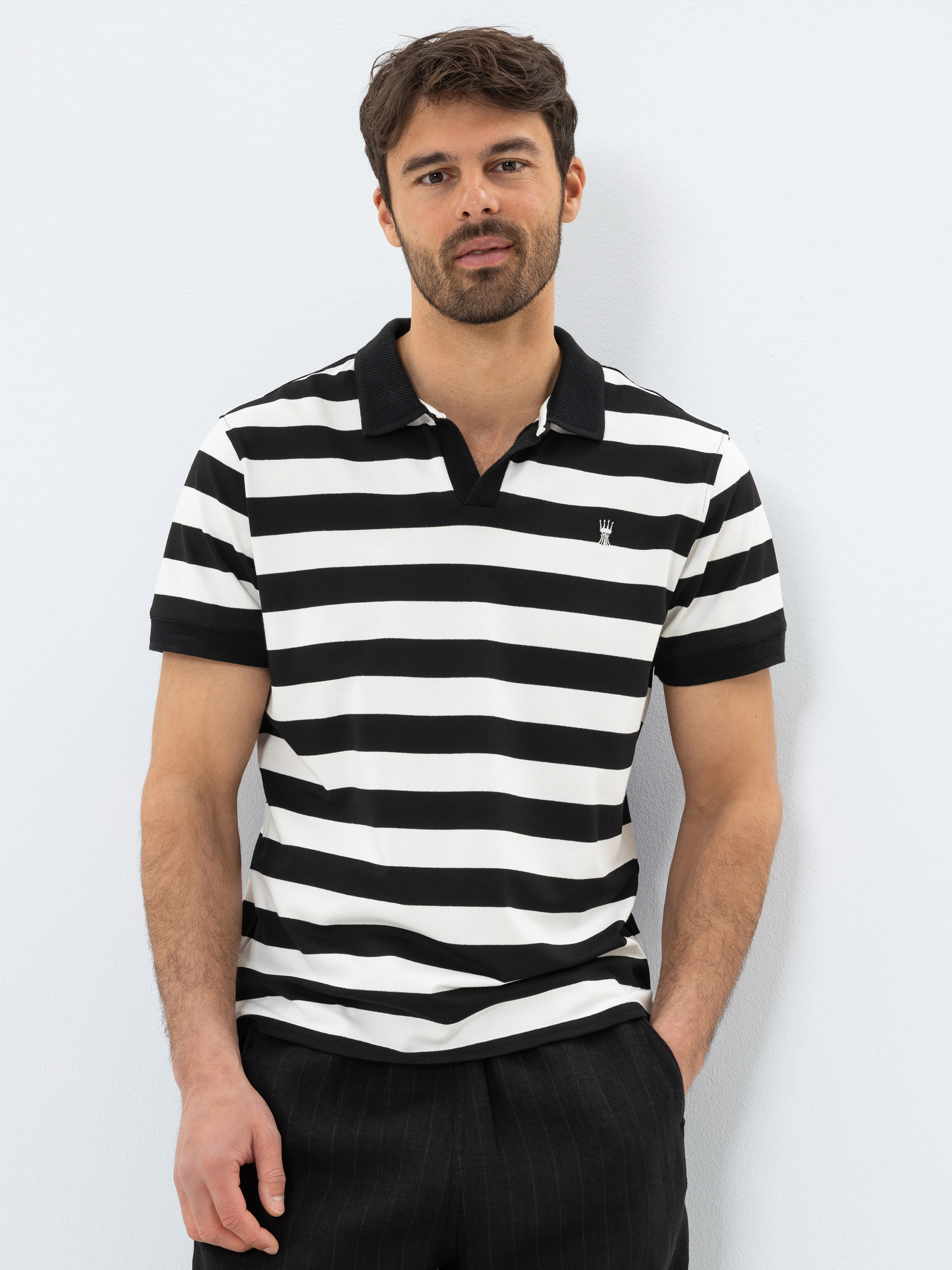 Ein Mann mit dunklem Haar und Bart trägt ein schwarz-weißes, horizontal gestreiftes Poloshirt mit einem kleinen Kronenlogo. Er steht vor einem schlichten hellen Hintergrund und hat die Hände in den Taschen.