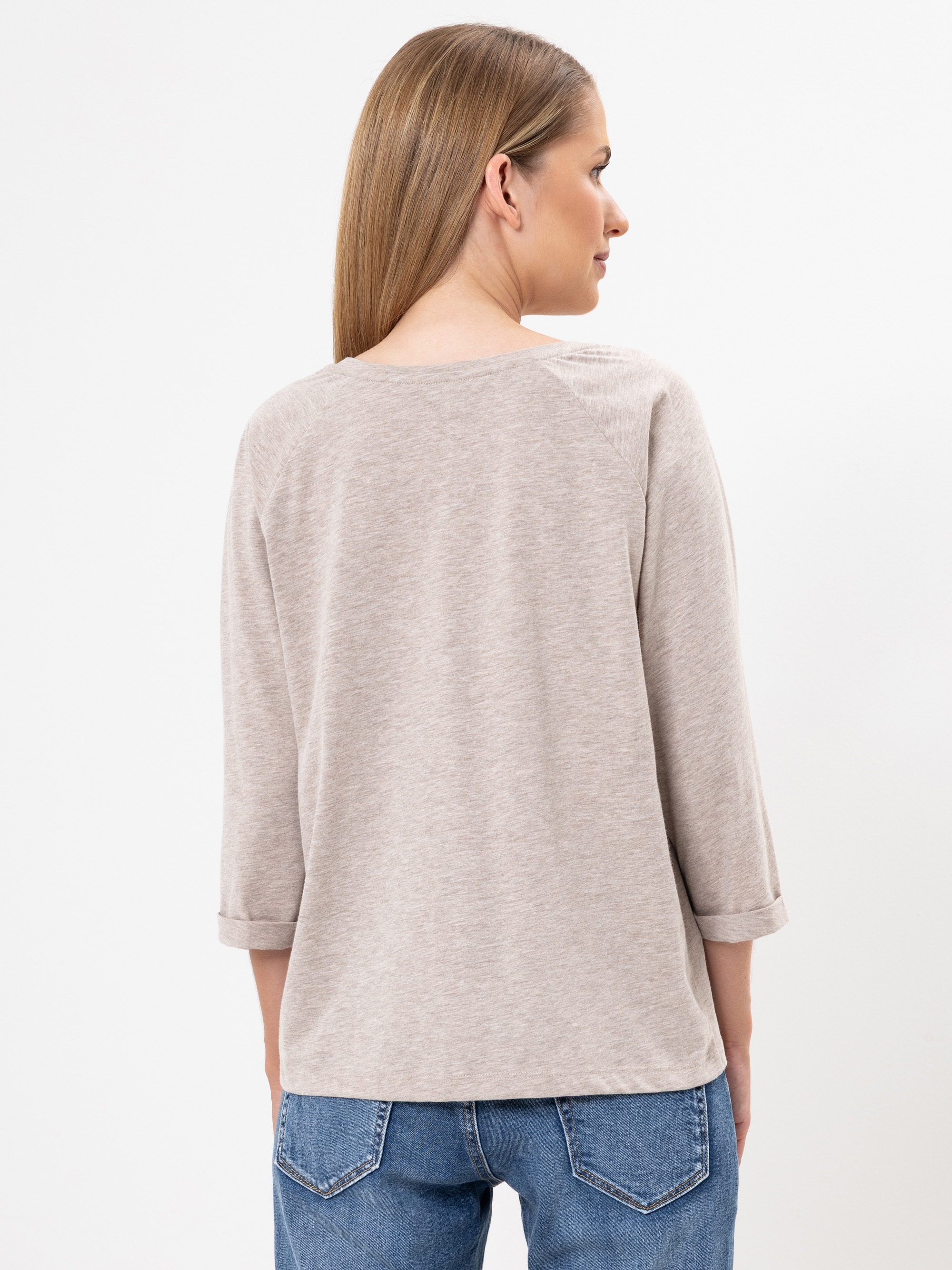 Eine Frau mit glattem, hellbraunem Haar steht vor einem einfarbig weißen Hintergrund. Sie trägt ein beigefarbenes, langärmeliges Oberteil mit Manschettenärmeln und blaue Jeans.